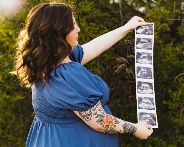 Maternity session in Indiana with pregnant woman holding ultrasound photos outdoors in natural setting.