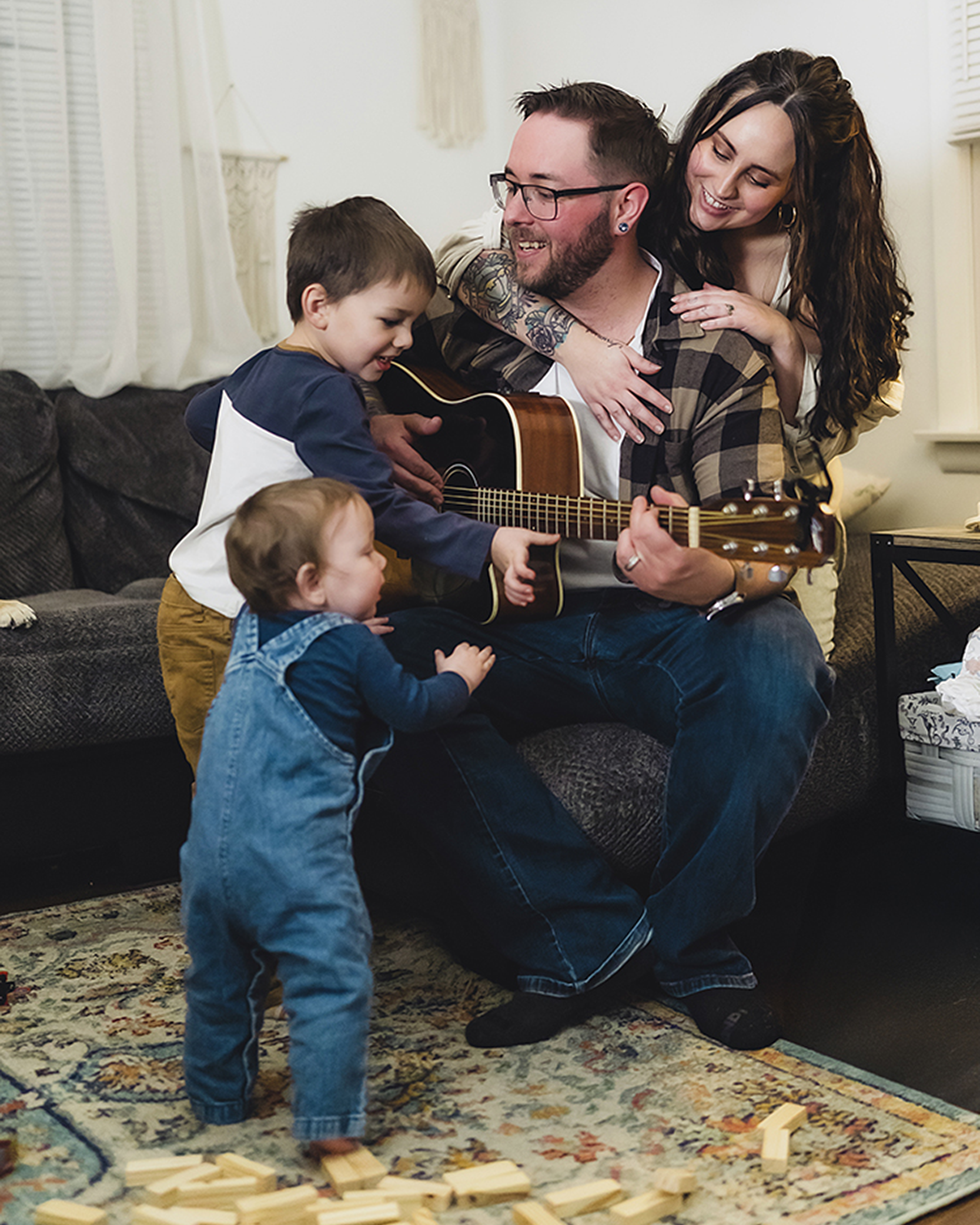 An in home photoshoot with a family of four enjoying time together in a cozy living room, with the father playing a guitar while the mother and two children smile and interact around him.