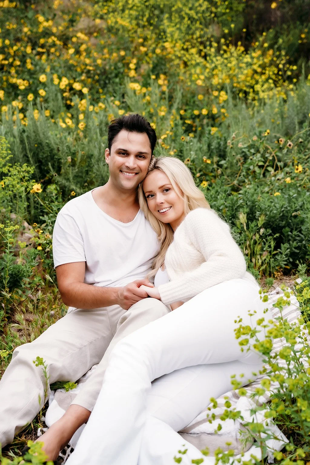 A happy couple sitting on a blanket in a field of yellow flower fields of Indiana, smiling at the camera.