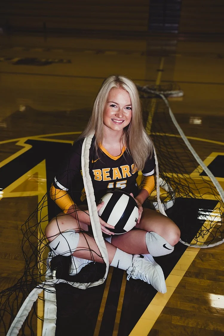High school senior volleyball player sitting on a gym floor holding a volleyball during a hobby-based senior portrait session.