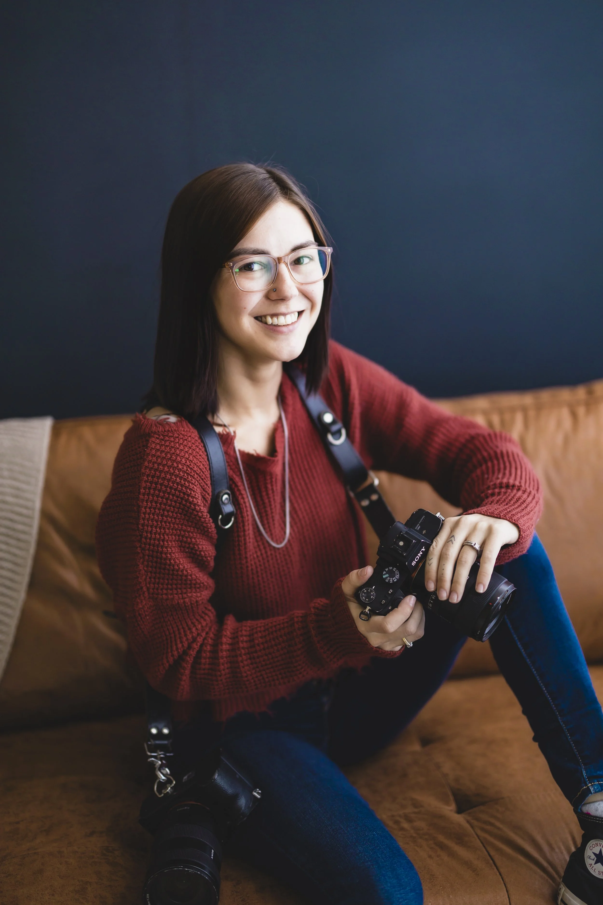 Richmond photographer headshot: woman with shoulder-length dark hair sitting on brown couch, holding Sony camera, smiling.
