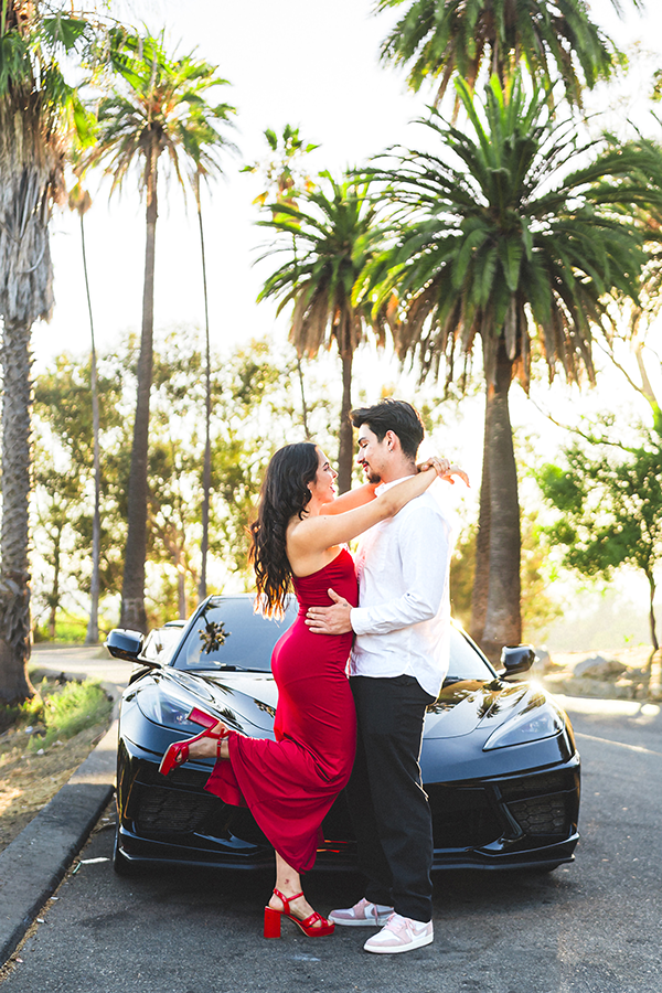 Couple in Elysian Park standing in front of a black sports car, embracing with palm trees and bright sunlight in the background.