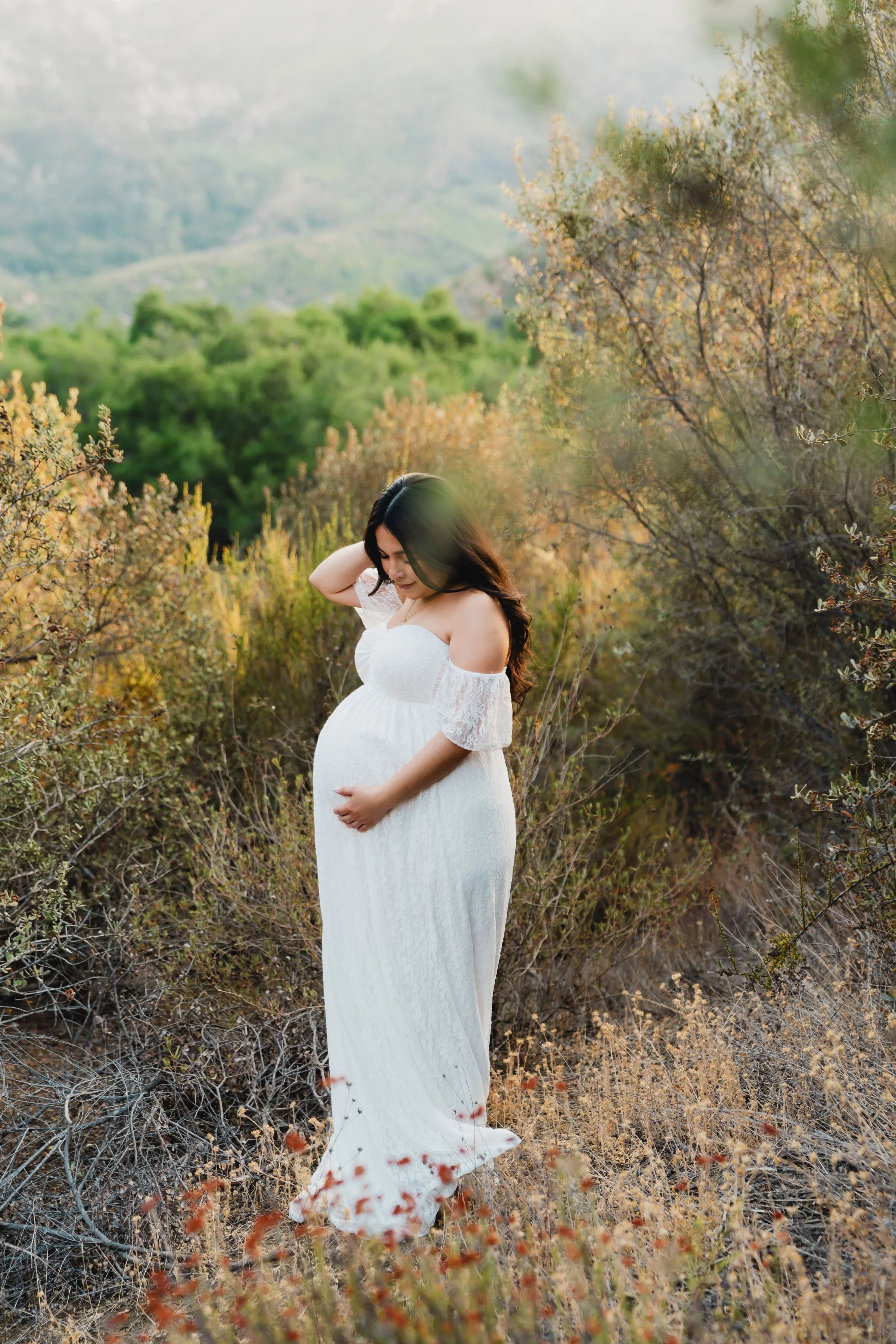 A pregnant woman in a white lace dress standing amidst dry bushes and greenery in a natural outdoor setting, gently holding her belly and looking down, with hills and trees in the background of scenic Indiana.