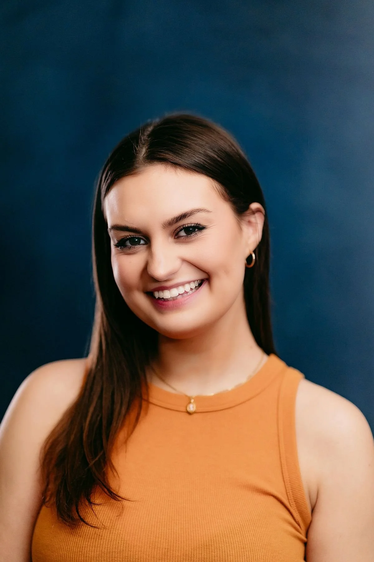 Yearbook style Senior portrait headshot of a young woman with long dark hair wearing an orange sleeveless top against a dark blue backdrop.