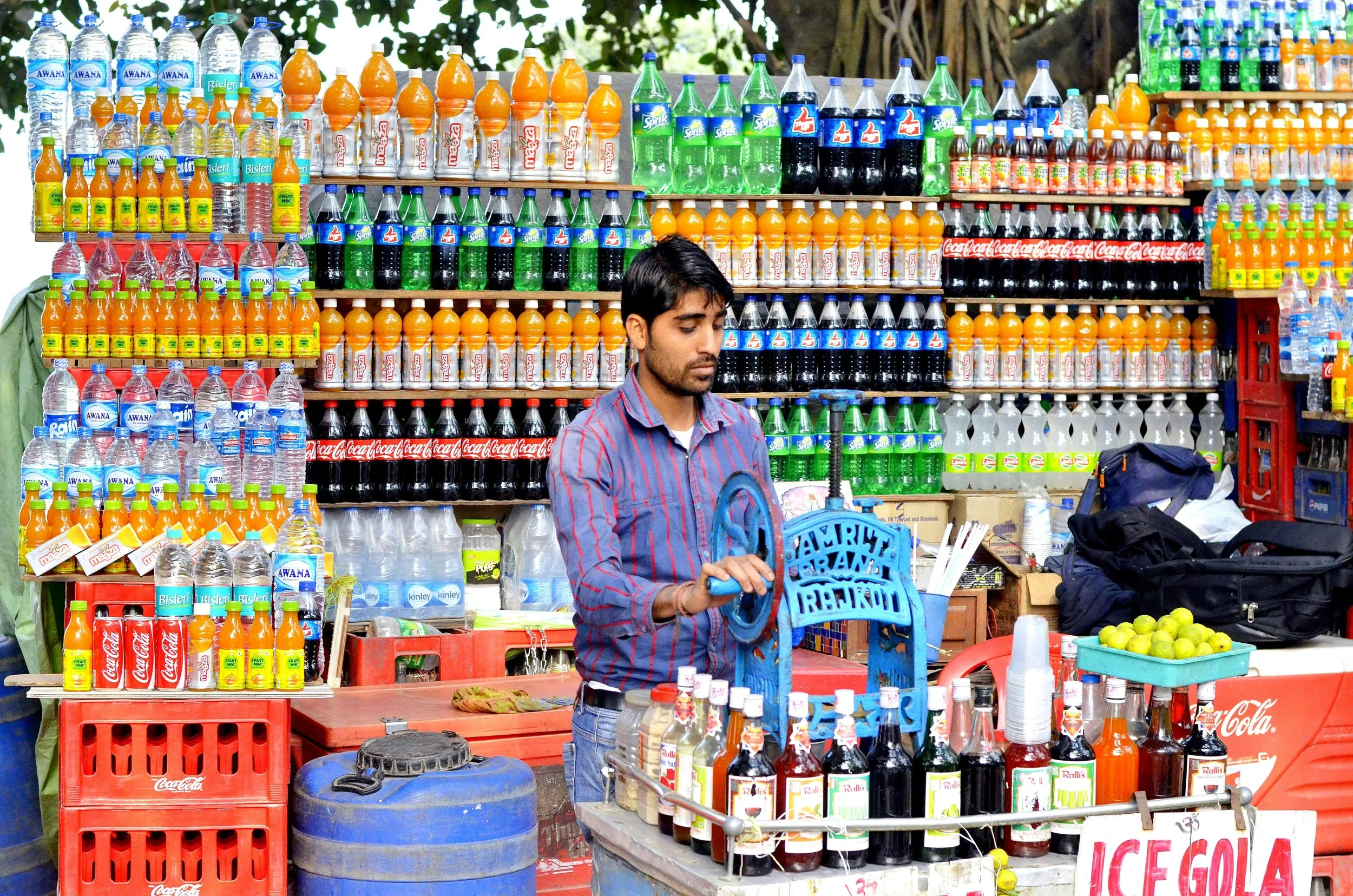 A man standing behind a street vendor's stall displaying bottles of soda and water. The stall has several shelves filled with colorful bottled drinks including Coke, Sprite, Fanta, and local brands, with some lemons on a tray nearby.