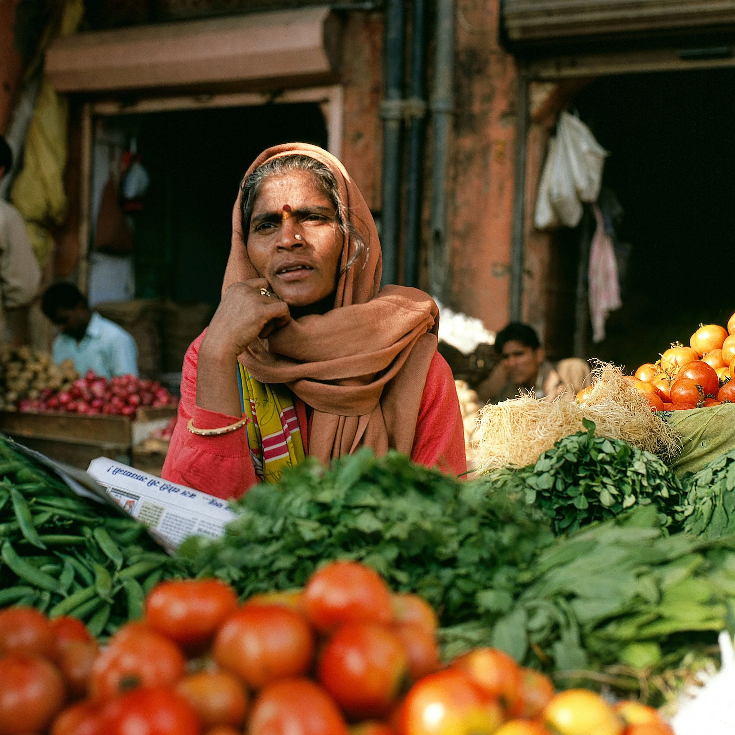A woman wearing a headscarf is at an outdoor market surrounded by fresh tomatoes and vegetables.