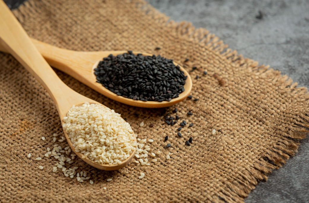 Two wooden spoons filled with sesame seeds, one with white seeds and the other with black seeds, placed on a woven brown fabric on a gray surface.