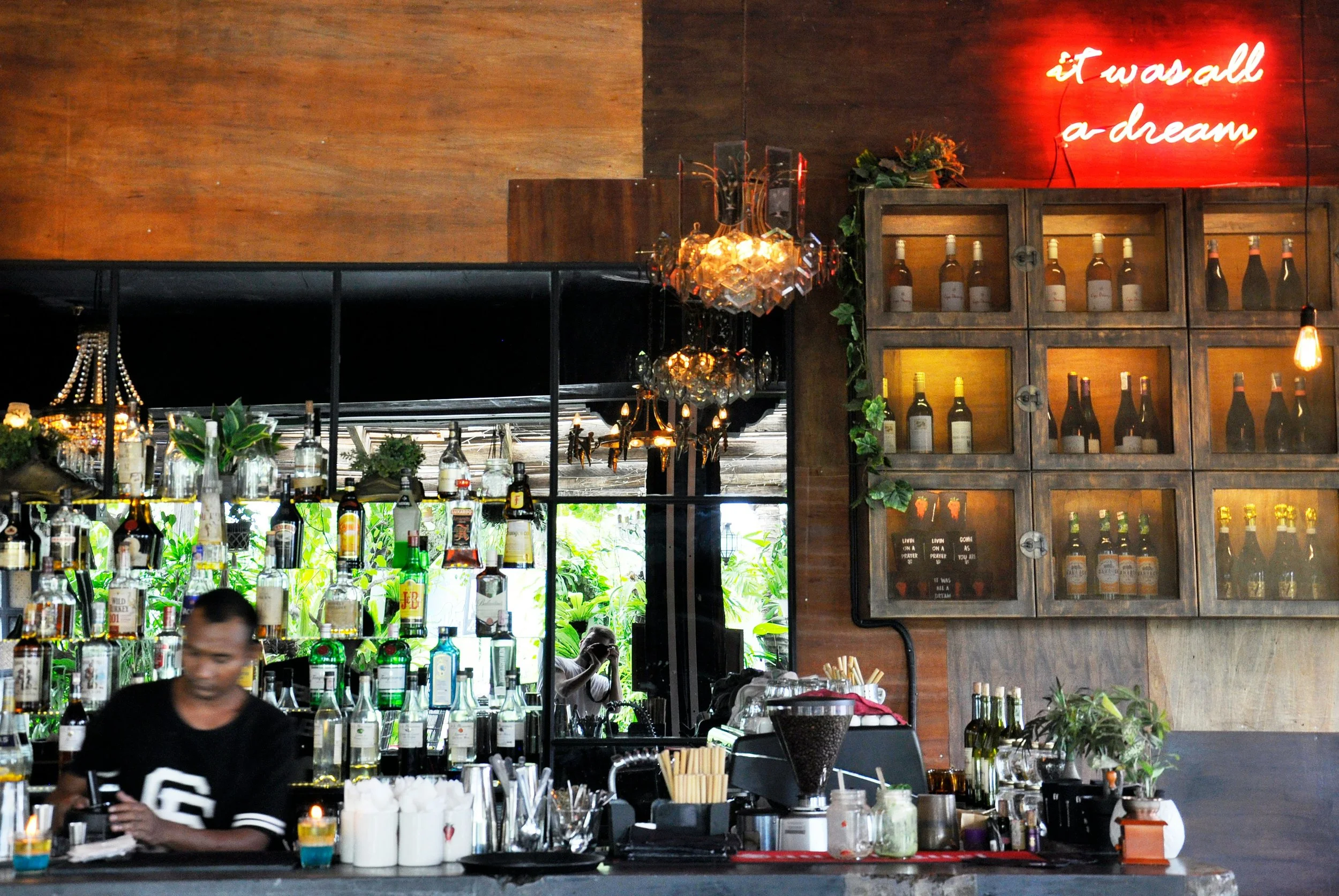 A bar with shelves of various alcohol bottles, a bartender in black shirt, and a decorative chandelier hanging from the ceiling. A neon sign on the wall reads 'it was all a dream'.