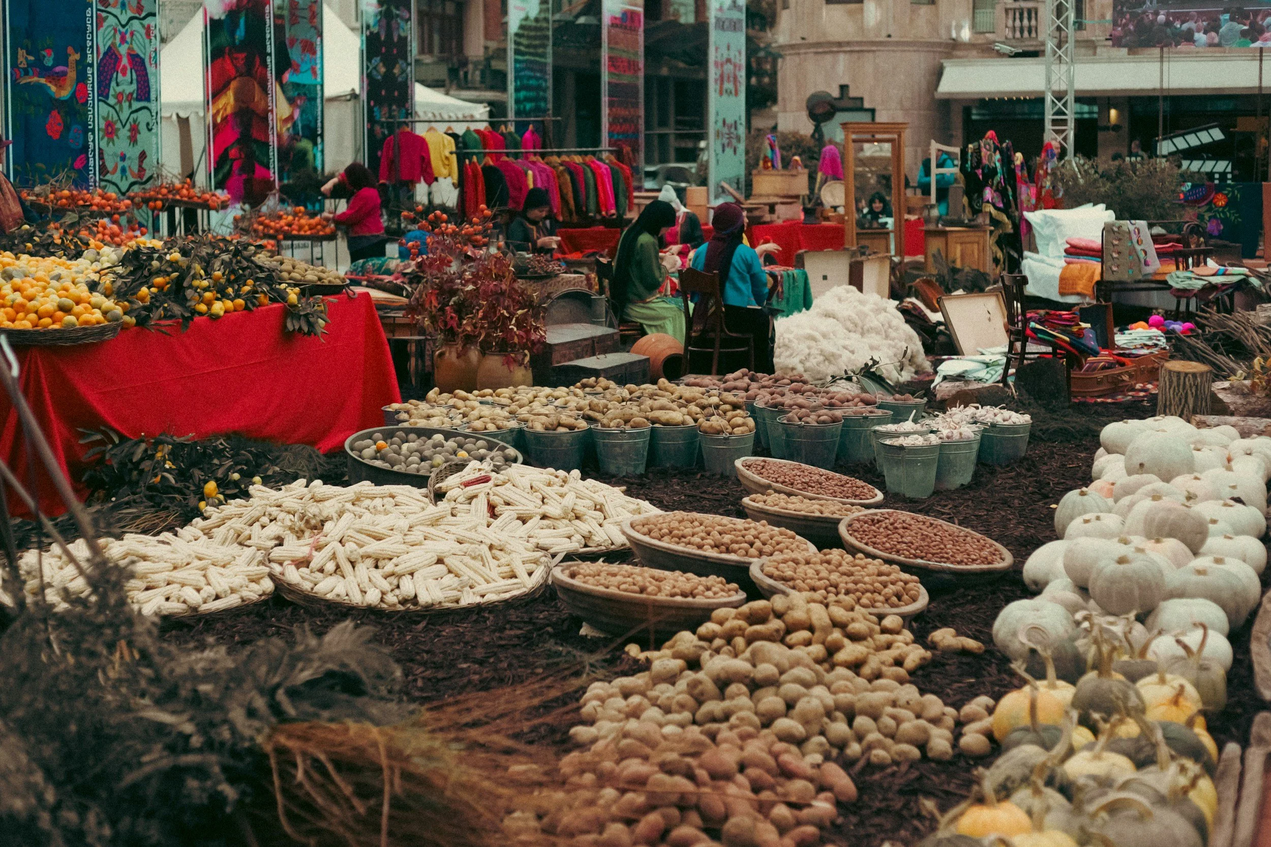 Outdoor market with various fruits, vegetables, and textiles displayed for sale, and vendors sitting behind their stalls.