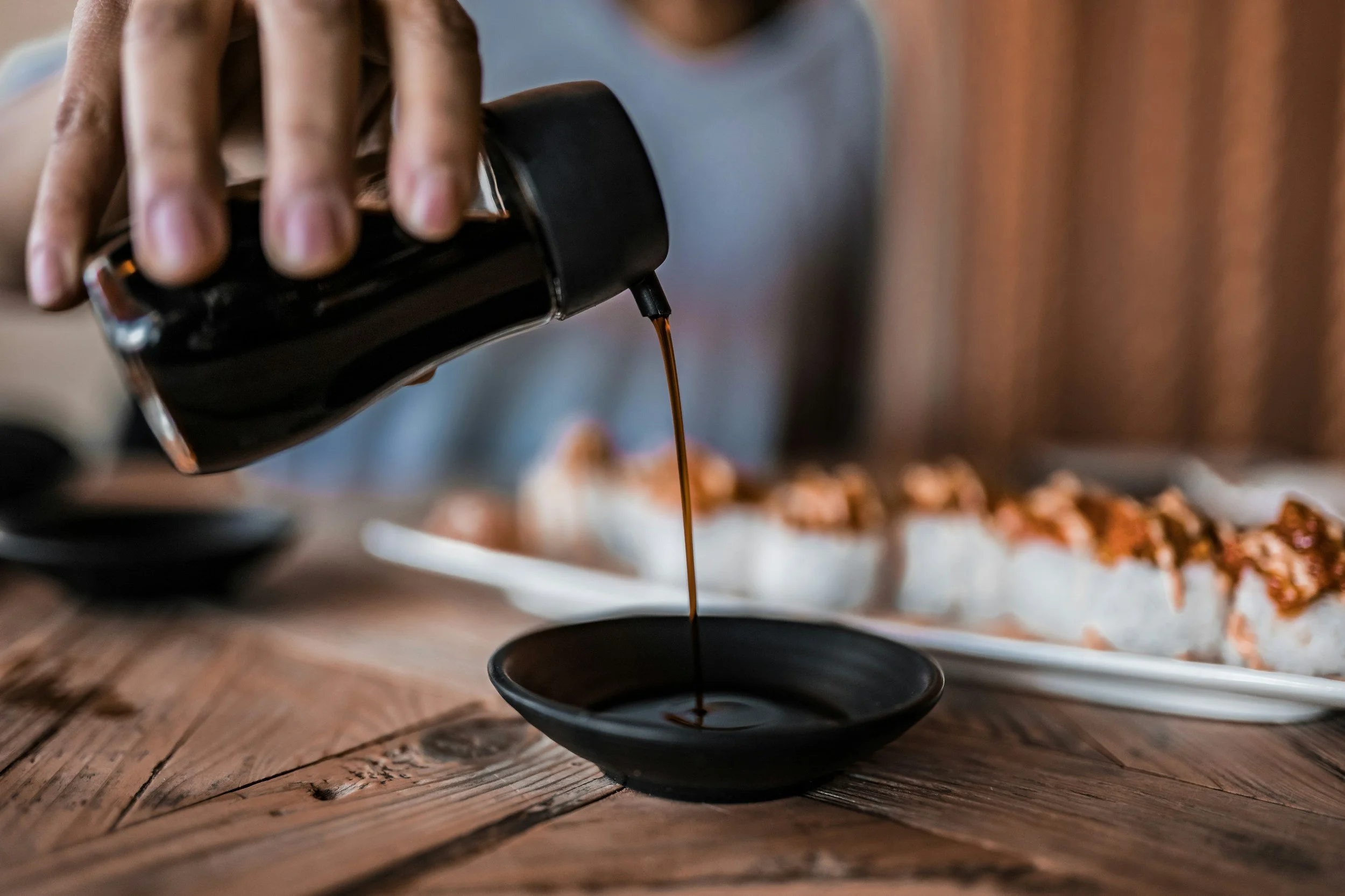 Person pouring soy sauce into a small black dish, with a tray of sushi in the background on a wooden table.