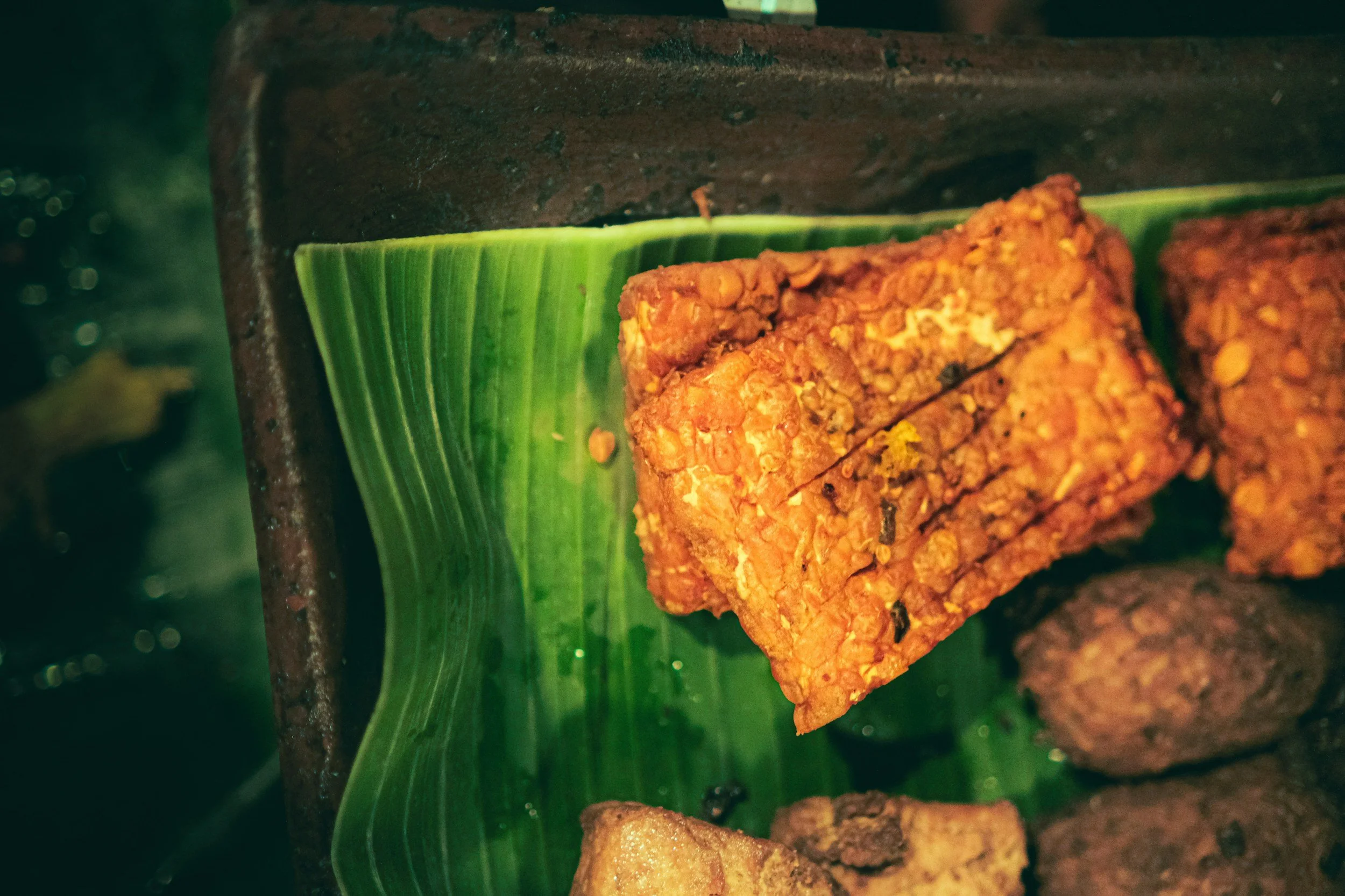 Fried snack items, possibly tofu or tempeh, on a banana leaf in a rustic setting.