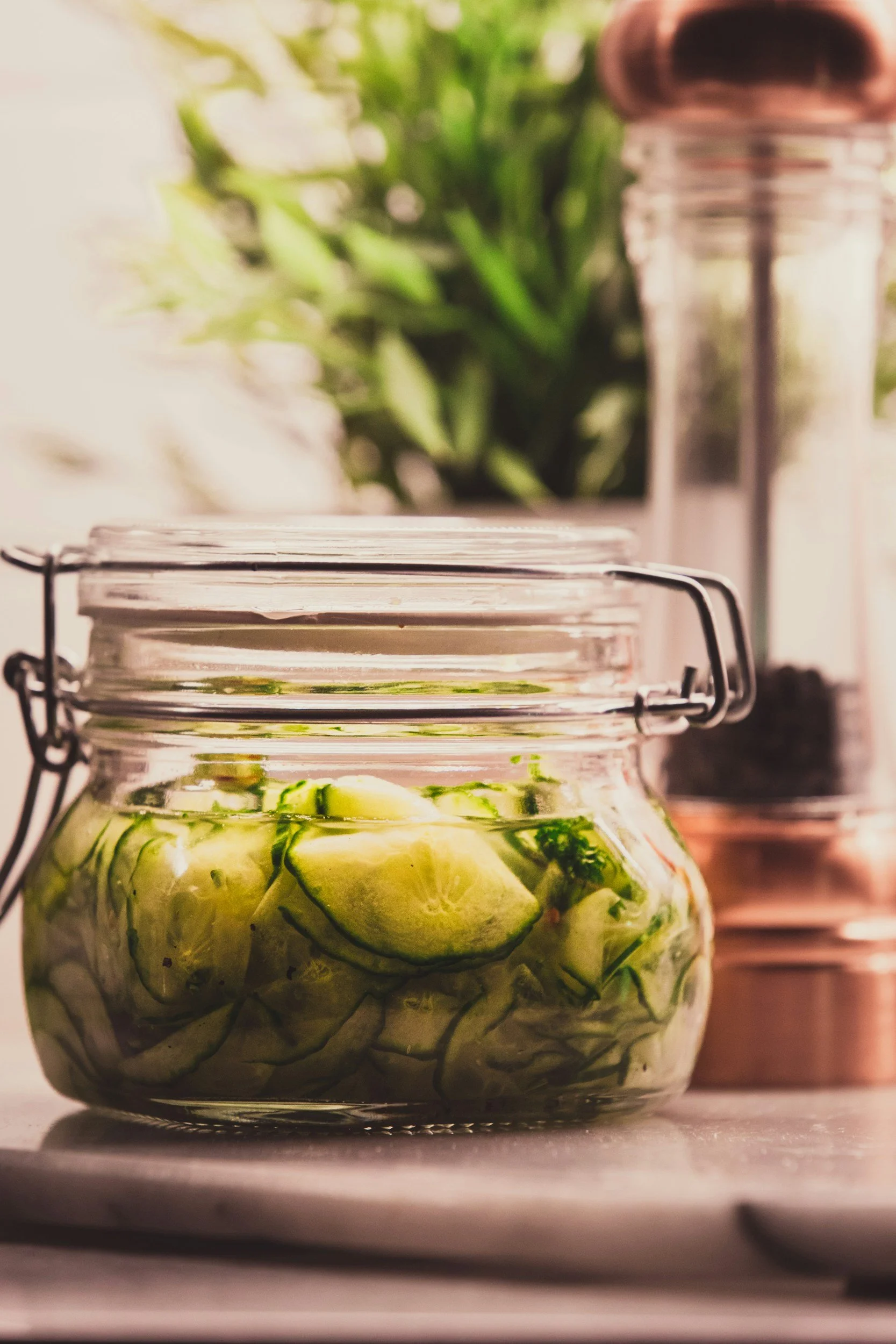 A glass jar filled with sliced cucumbers, ready for pickling, placed on a white surface with a blurred background of green leaves and kitchen spices.
