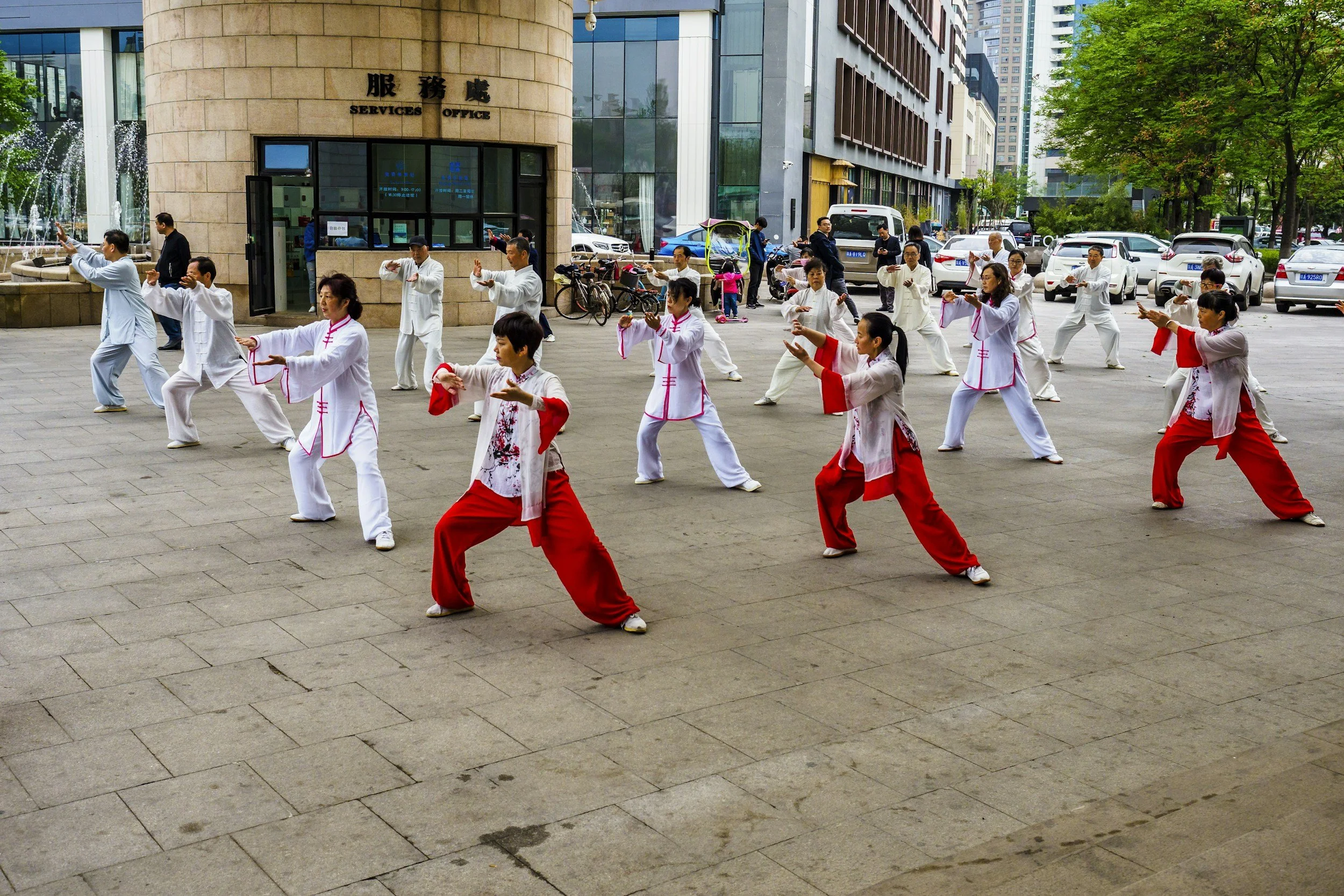 Group of people practicing Tai Chi or martial arts in an outdoor urban plaza, with some wearing traditional Chinese clothing in red and white, and others in white uniforms.