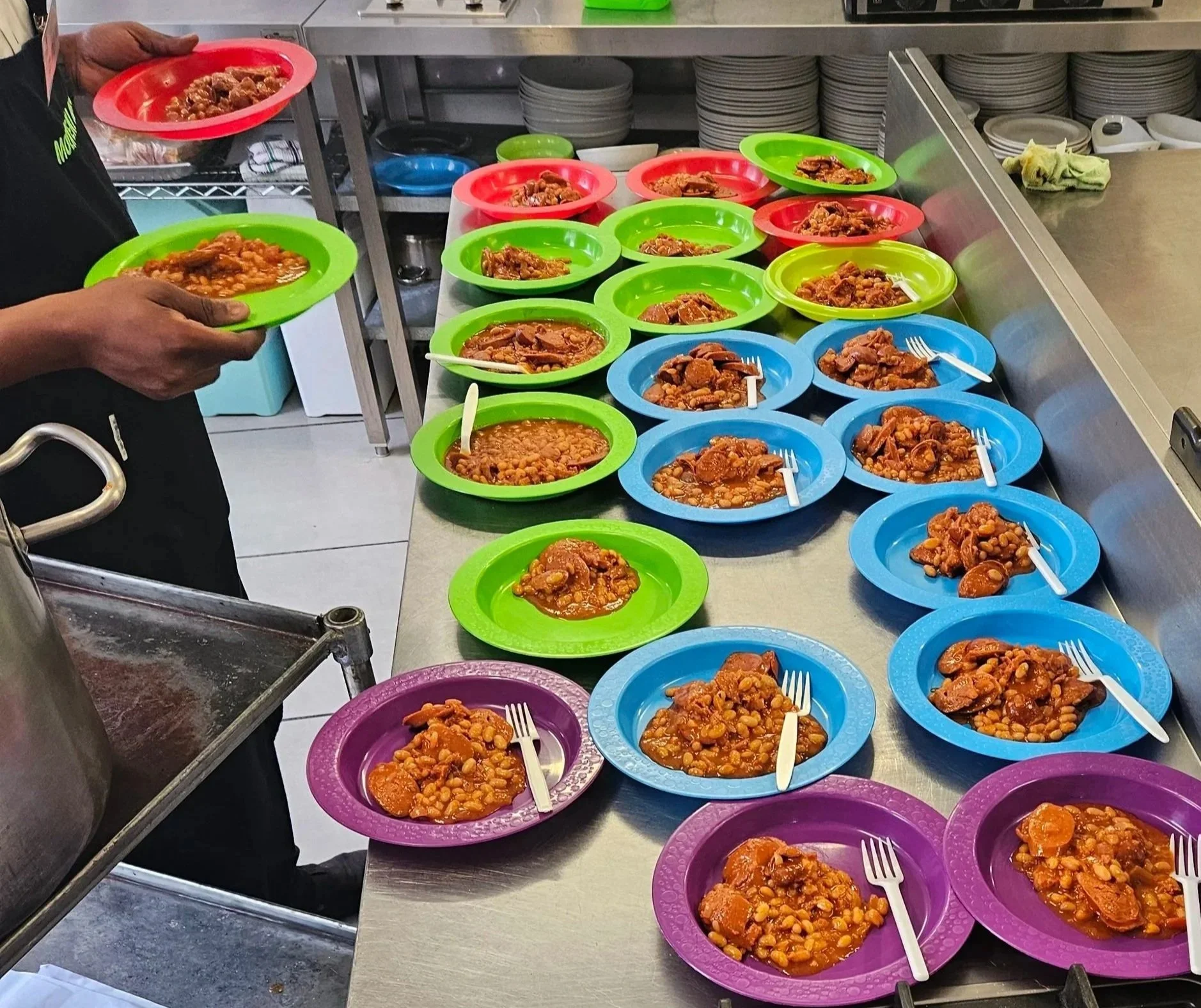 Display of multiple plates with beans and meat stew, colorful plates including purple, blue, green, and pink, on a stainless steel counter in a kitchen or cafeteria setting.