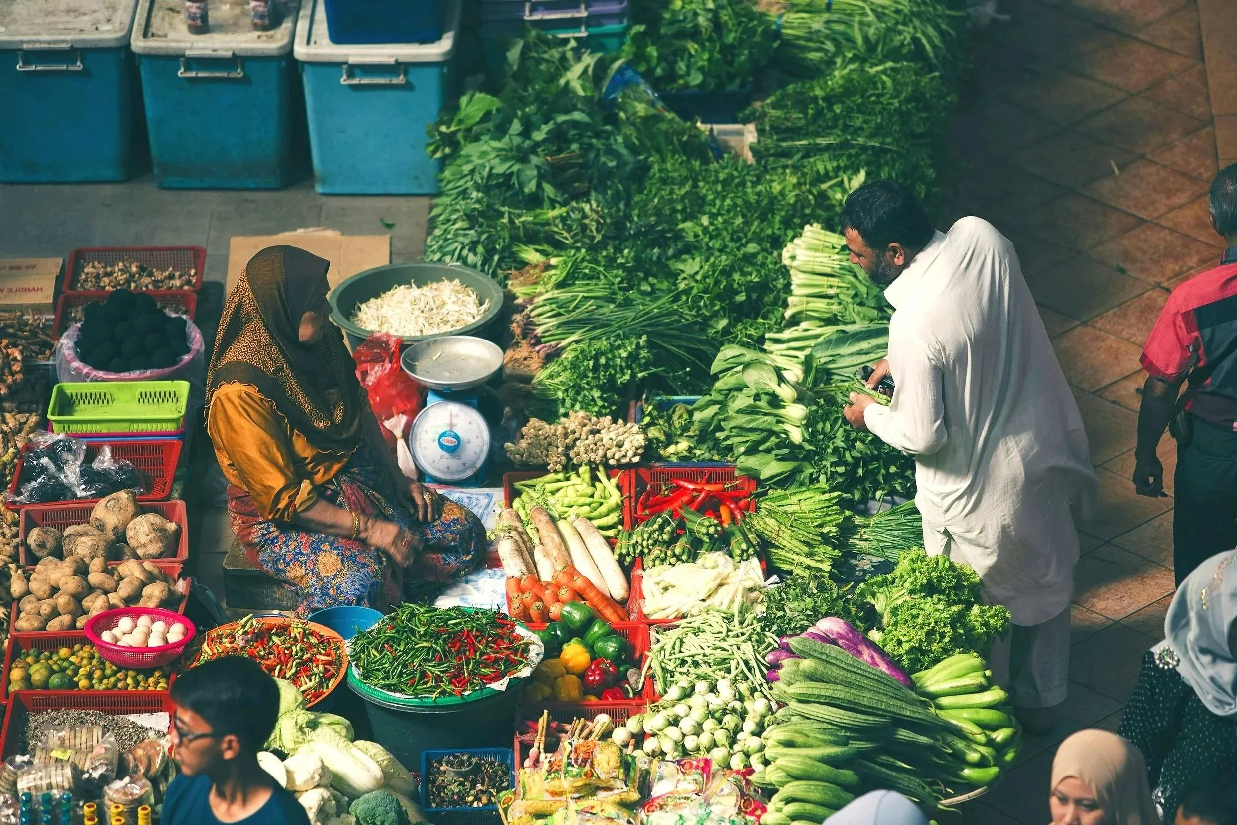 A busy outdoor market stall filled with fresh vegetables, leafy greens, and other produce. Several people are shopping, including a woman sitting among the vegetables and a man examining the produce. Large baskets and containers of vegetables such as carrots, radishes, potatoes, and greens are visible.