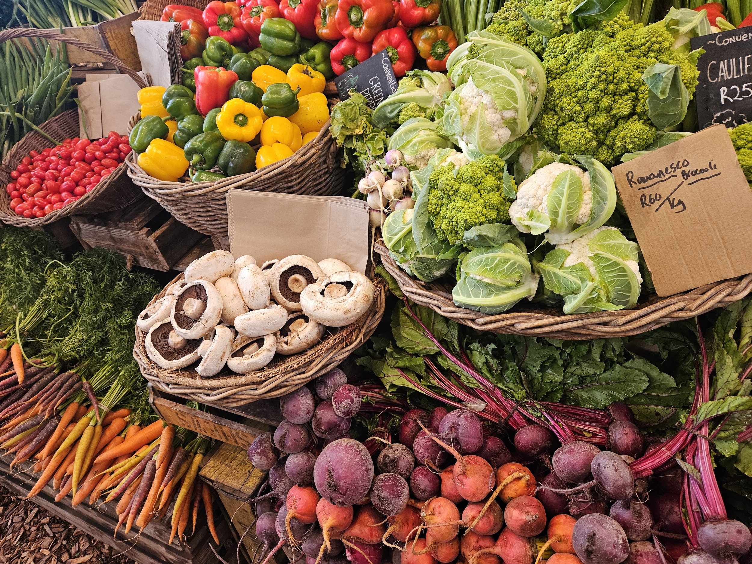 Various fresh vegetables including cherry tomatoes, yellow and green bell peppers, cauliflower, broccoli, radishes, mushrooms, beets, and carrots displayed in baskets at a market.