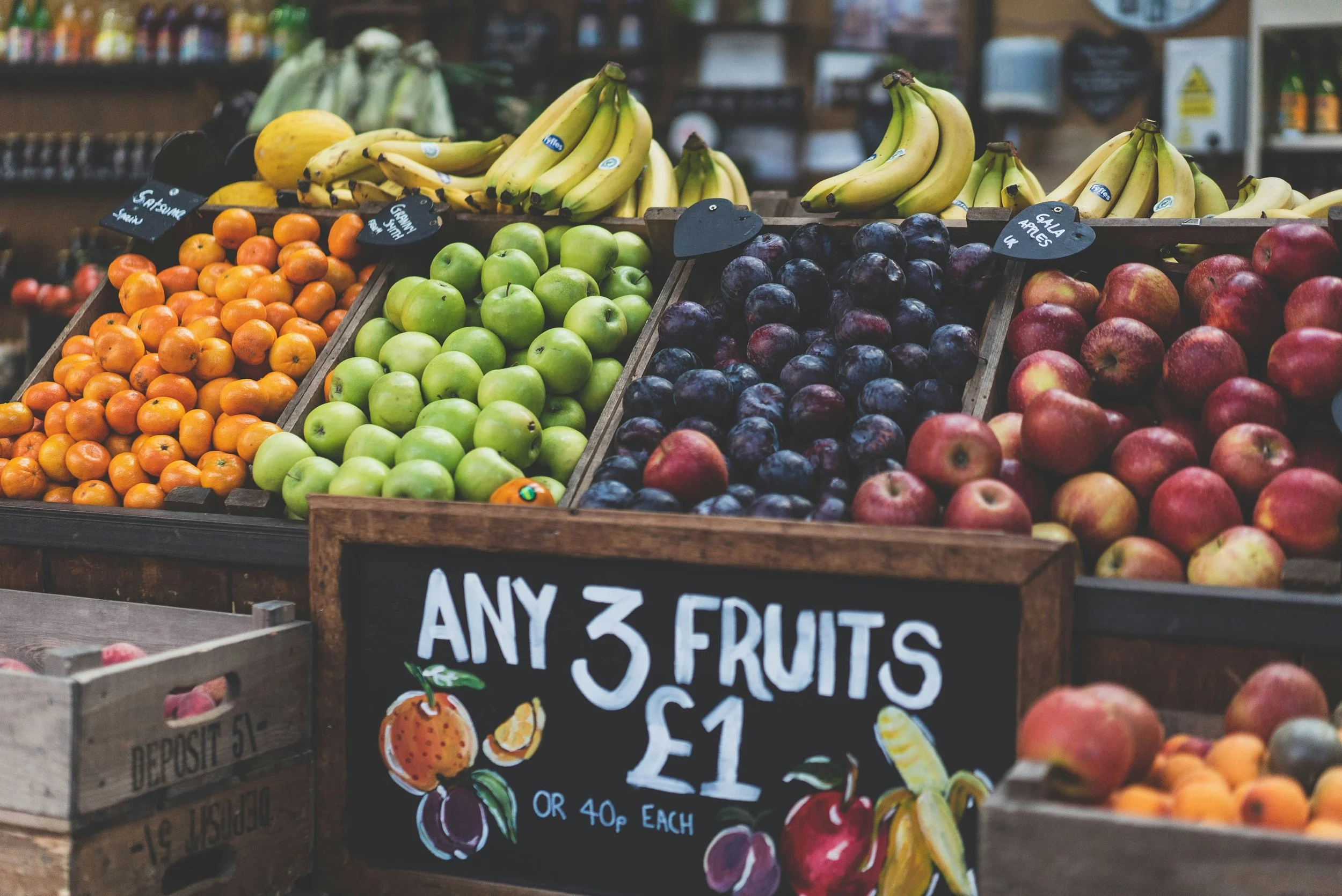 A display of various fresh fruits including oranges, green apples, black plums, bananas, and red apples at a market stand, with a sign advertising any 3 fruits for $1 or each for 40 cents.