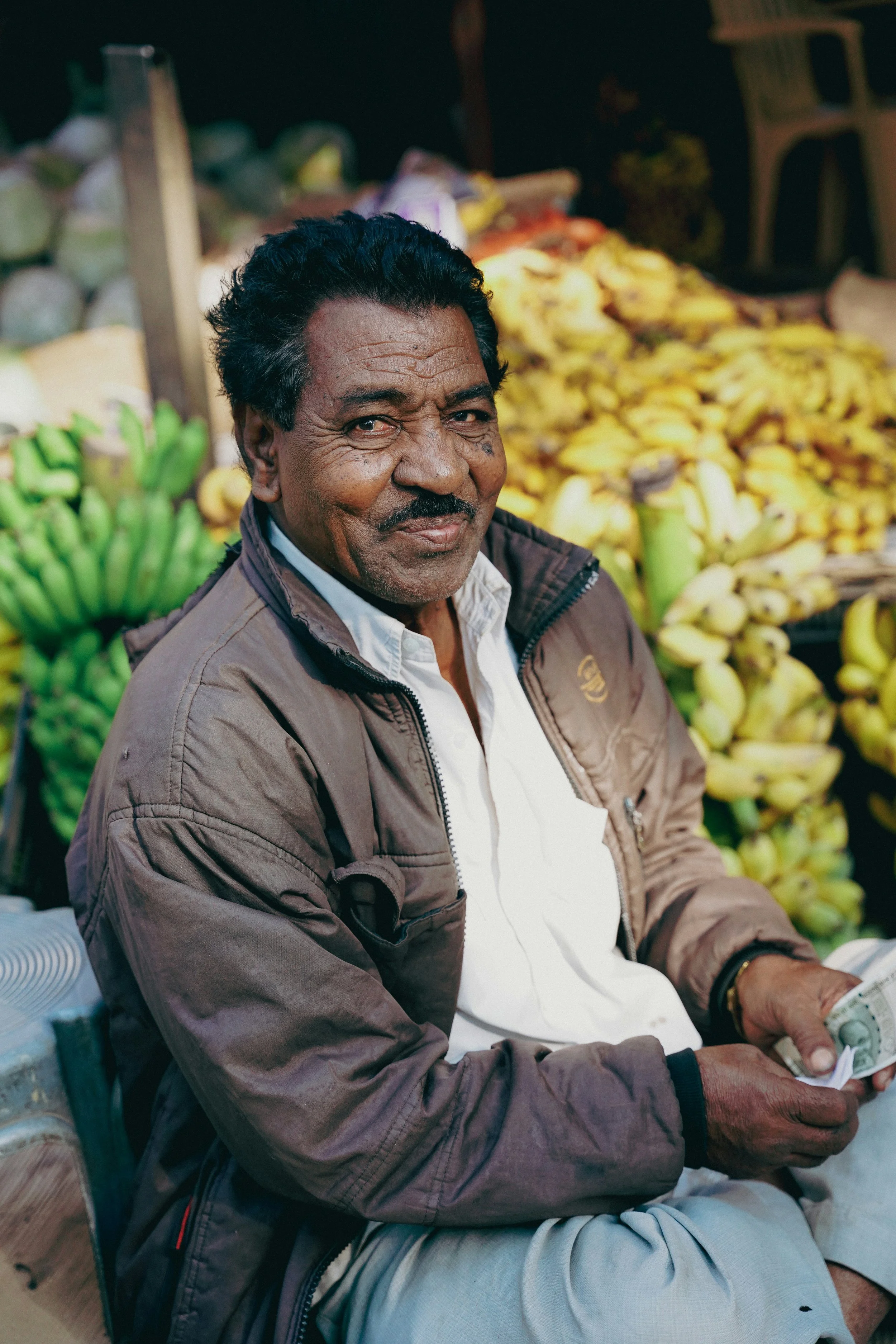 Man sitting at a fruit stand with a stack of bananas behind him, holding cash and smiling.