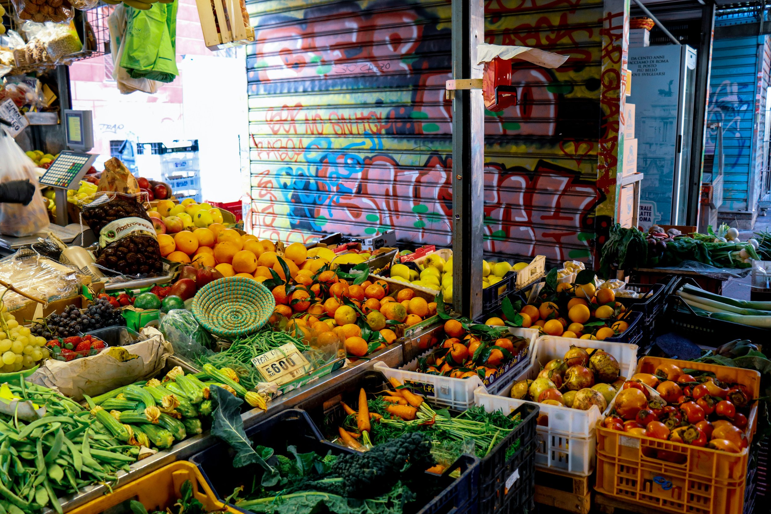 A market stall with a variety of fresh fruits and vegetables, including oranges, grapes, strawberries, carrots, and leafy greens, with graffiti-covered shutter in the background.