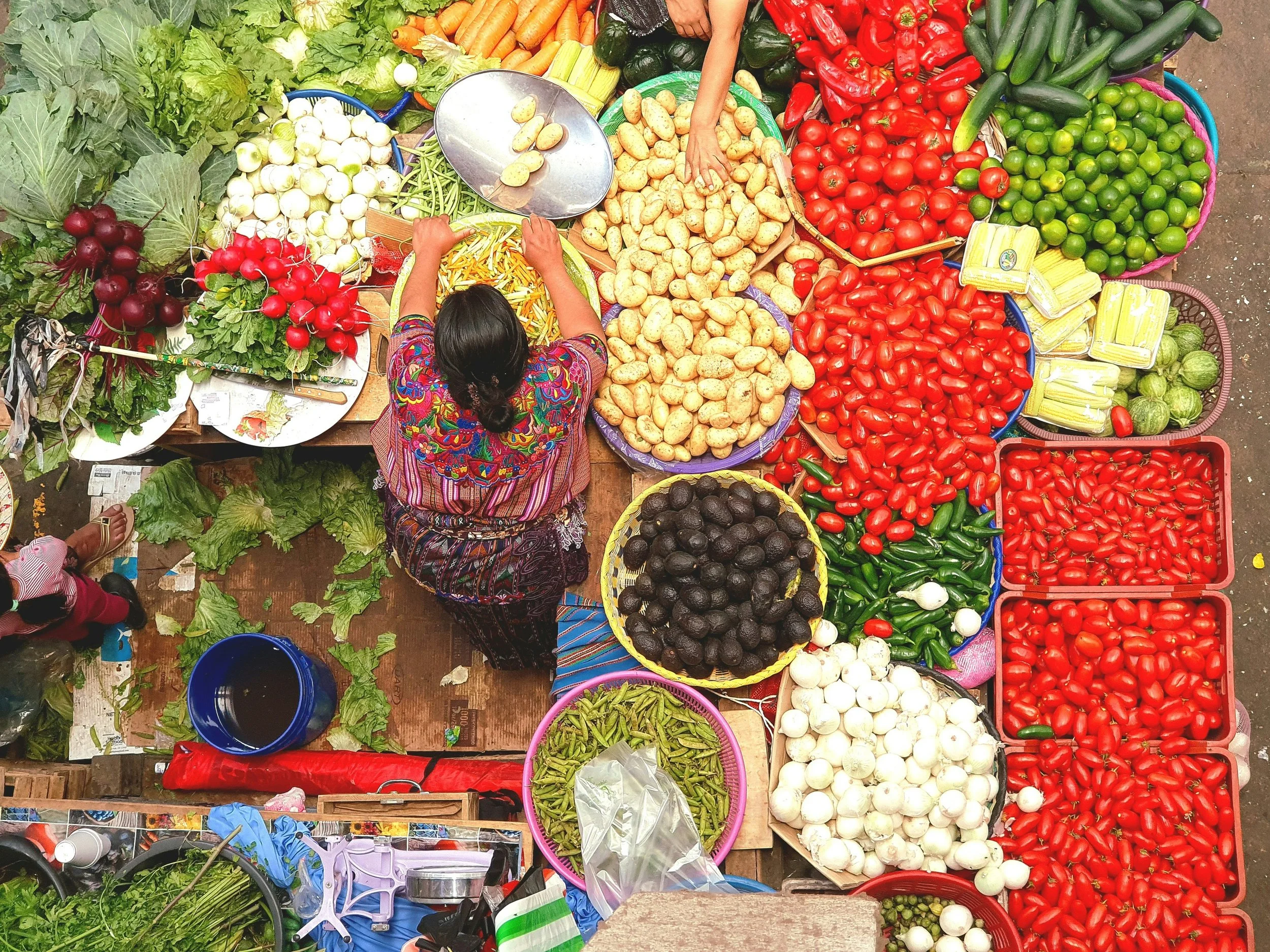 A street market vendor displays a variety of fresh vegetables in colorful baskets including radishes, tomatoes, zucchini, potatoes, onions, and peppers, with a vendor arranging produce. A person is seen from above, shopping among the vegetables.