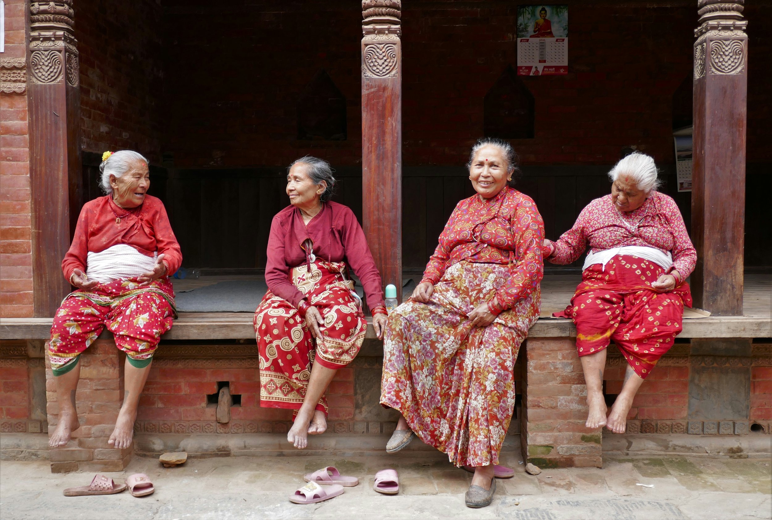 Four elderly women sitting on a porch in traditional red and floral-patterned clothing, smiling and chatting with each other.