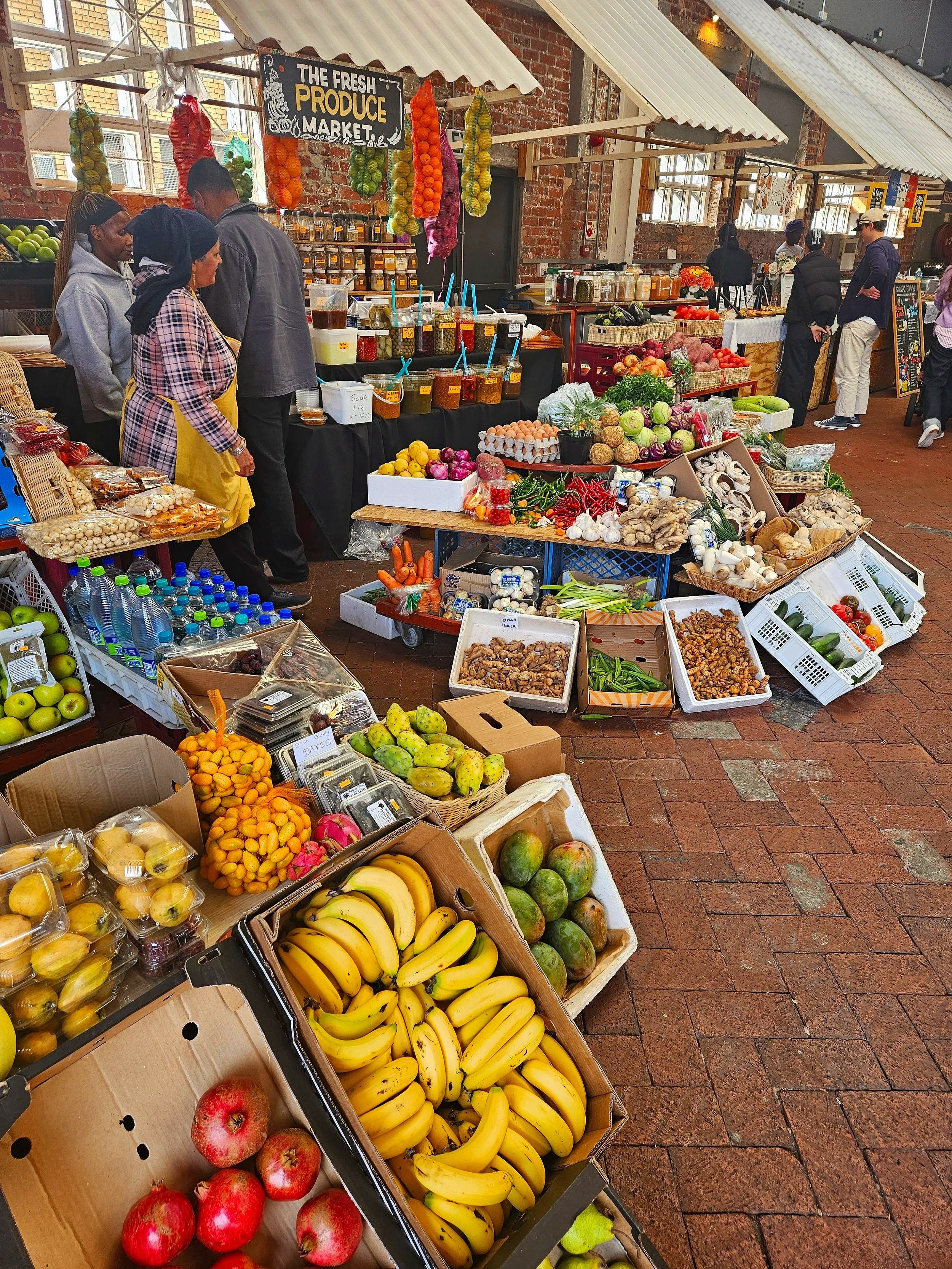 Market stall displaying fresh produce such as bananas, pomegranates, apples, garlic, carrots, and various vegetables, under a sign that reads 'The Fresh Produce Market' with customers shopping in an industrial-style market.