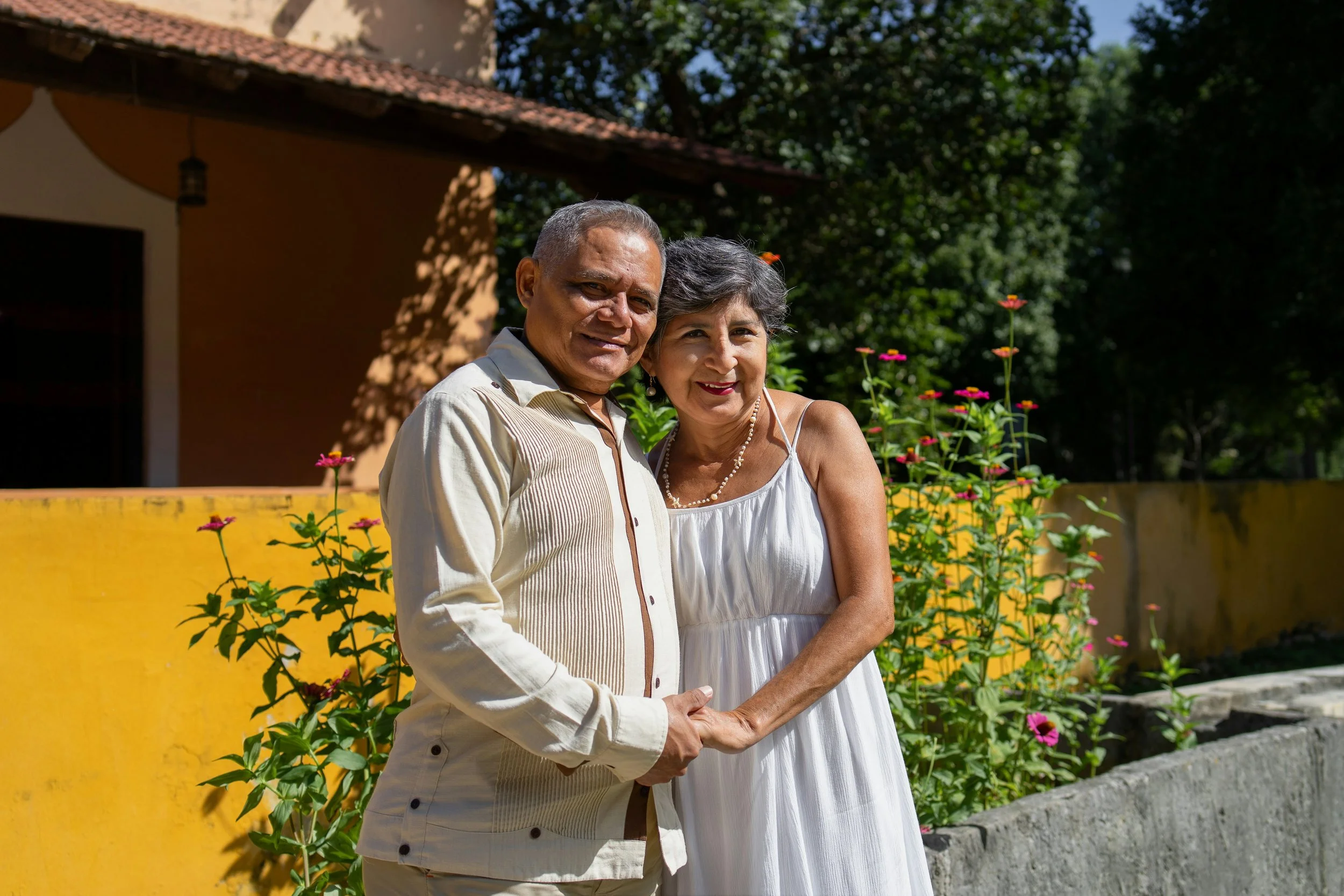 Older couple standing outdoors in front of a yellow wall with pink flowers, holding hands and smiling.