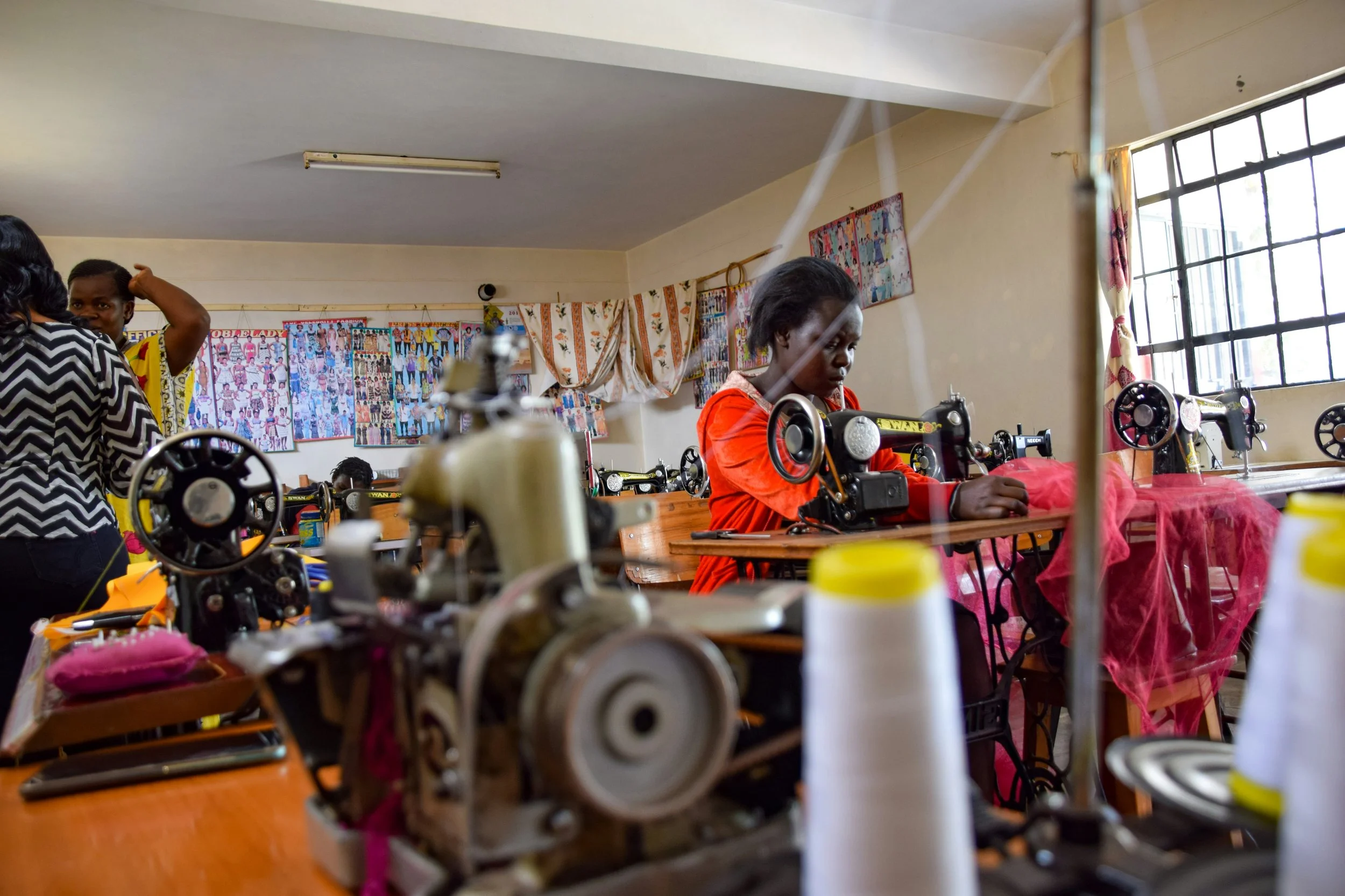 Young girl working with a sewing machine in a classroom, surrounded by fabric and sewing supplies, with colorful posters on the wall and a large window letting in natural light.