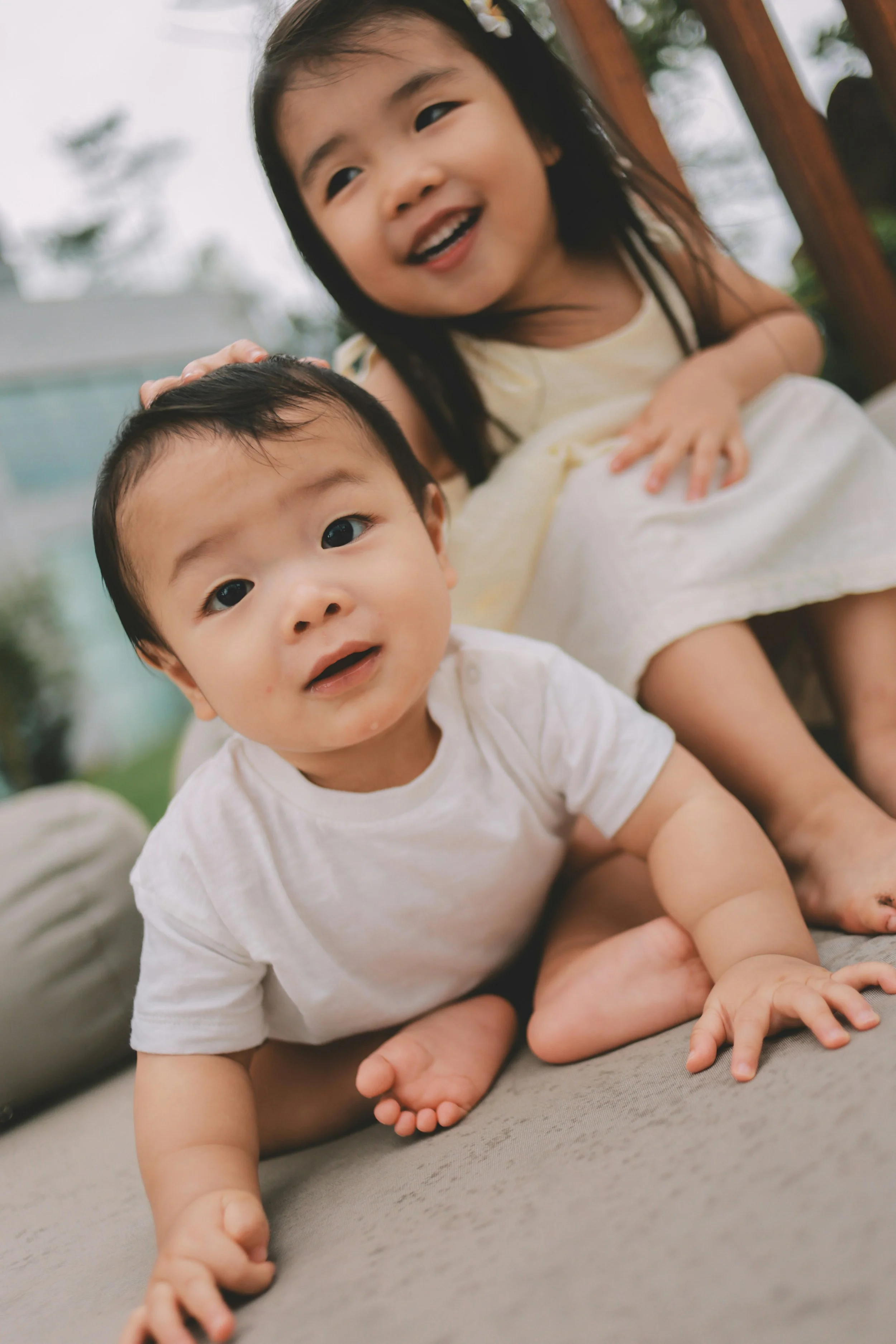 Two young children, a baby and a girl, playing on a couch, smiling and looking at the camera.
