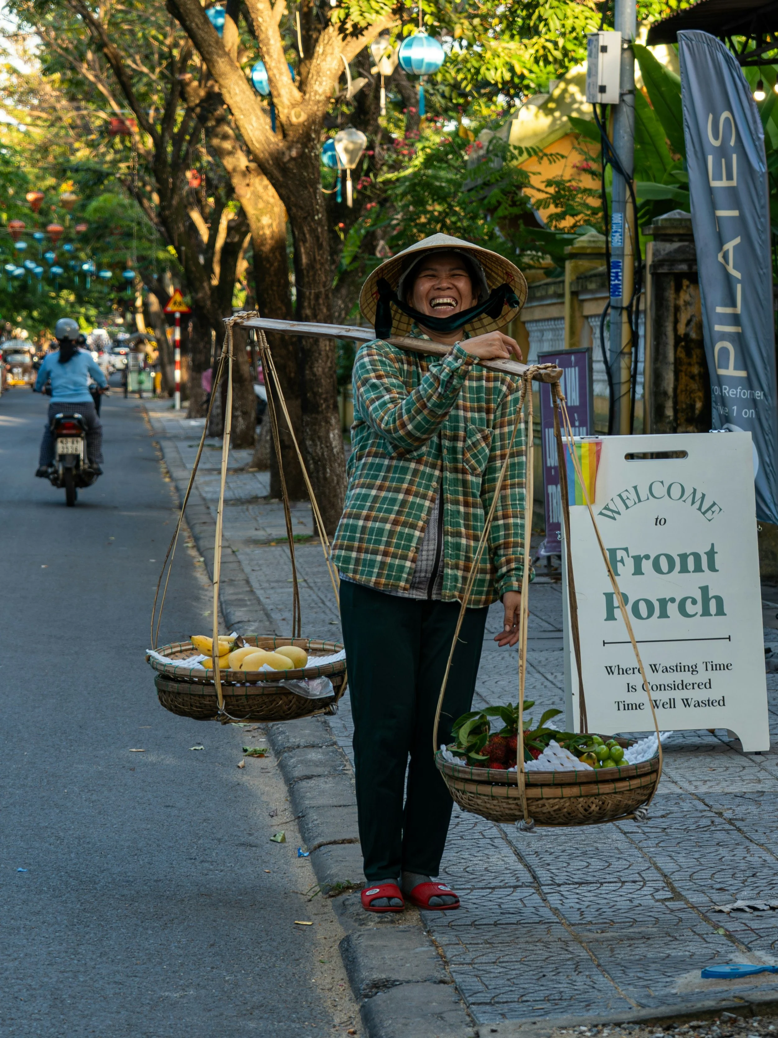 A street vendor woman wearing a traditional Vietnamese conical hat, smiling and carrying baskets of fruits including bananas and apples on a balance pole. Behind her, there is a sign that reads 'WELCOME to Front Porch,' and she is standing on a sidewalk next to a tree with hanging lanterns, with a motorbike and a person riding it visible in the background.