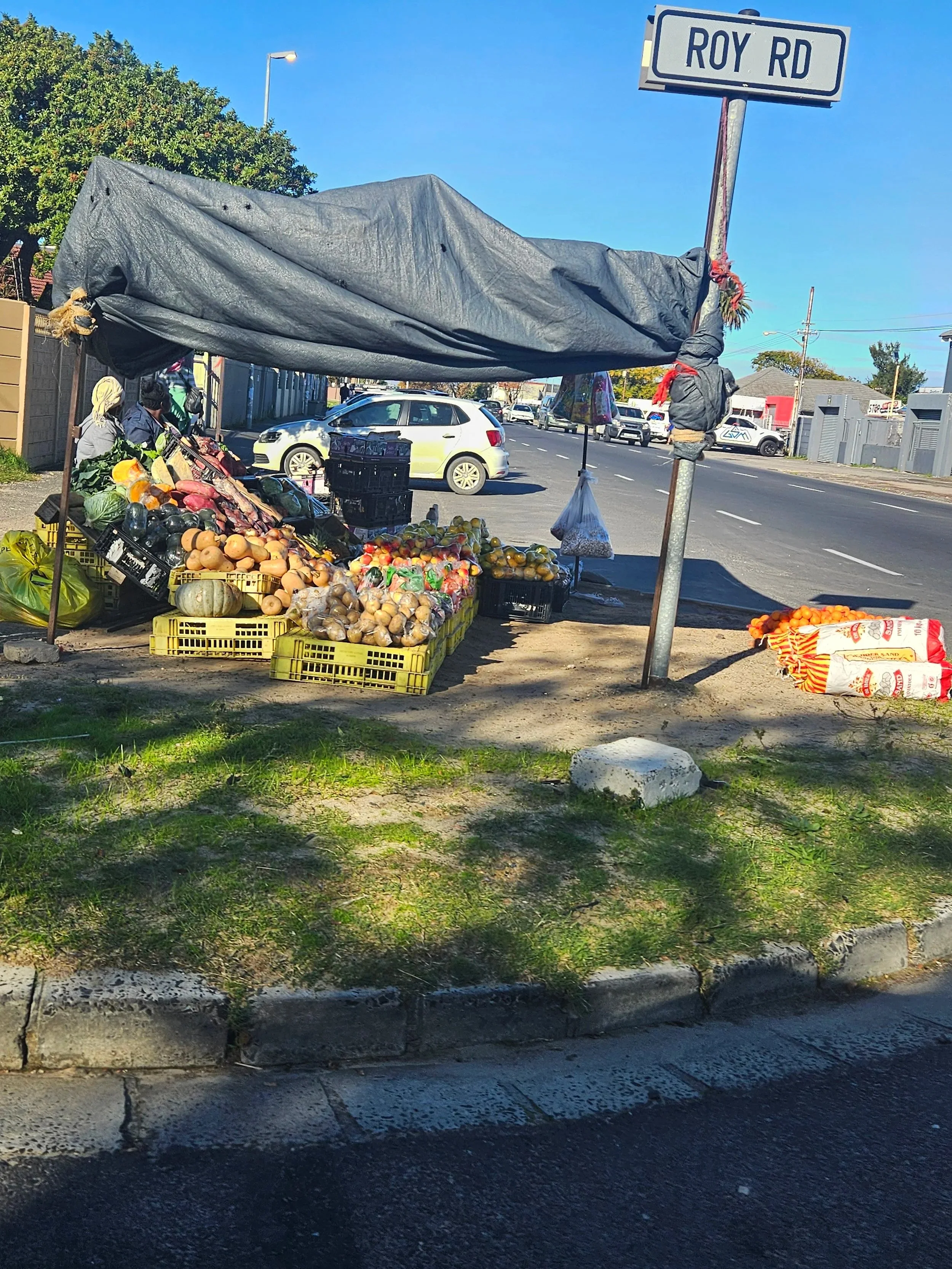 A street market stall with fruits and vegetables under a makeshift canopy on the sidewalk, next to a street sign that reads 'Roy Rd'. Cars are parked in the background on a sunny day with a blue sky.
