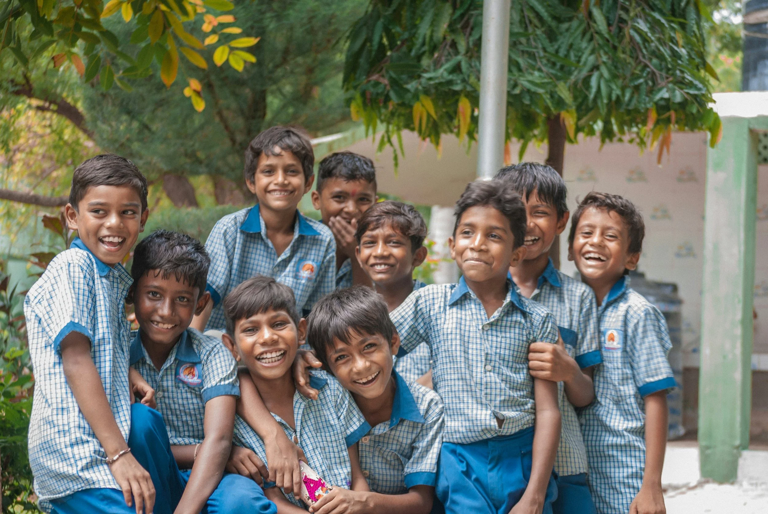 A group of smiling young boys in blue school uniforms standing together outdoors among trees.