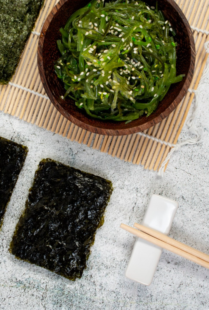Seaweed salad topped with sesame seeds in a wooden bowl, with sheets of nori seaweed and chopsticks on a white surface.