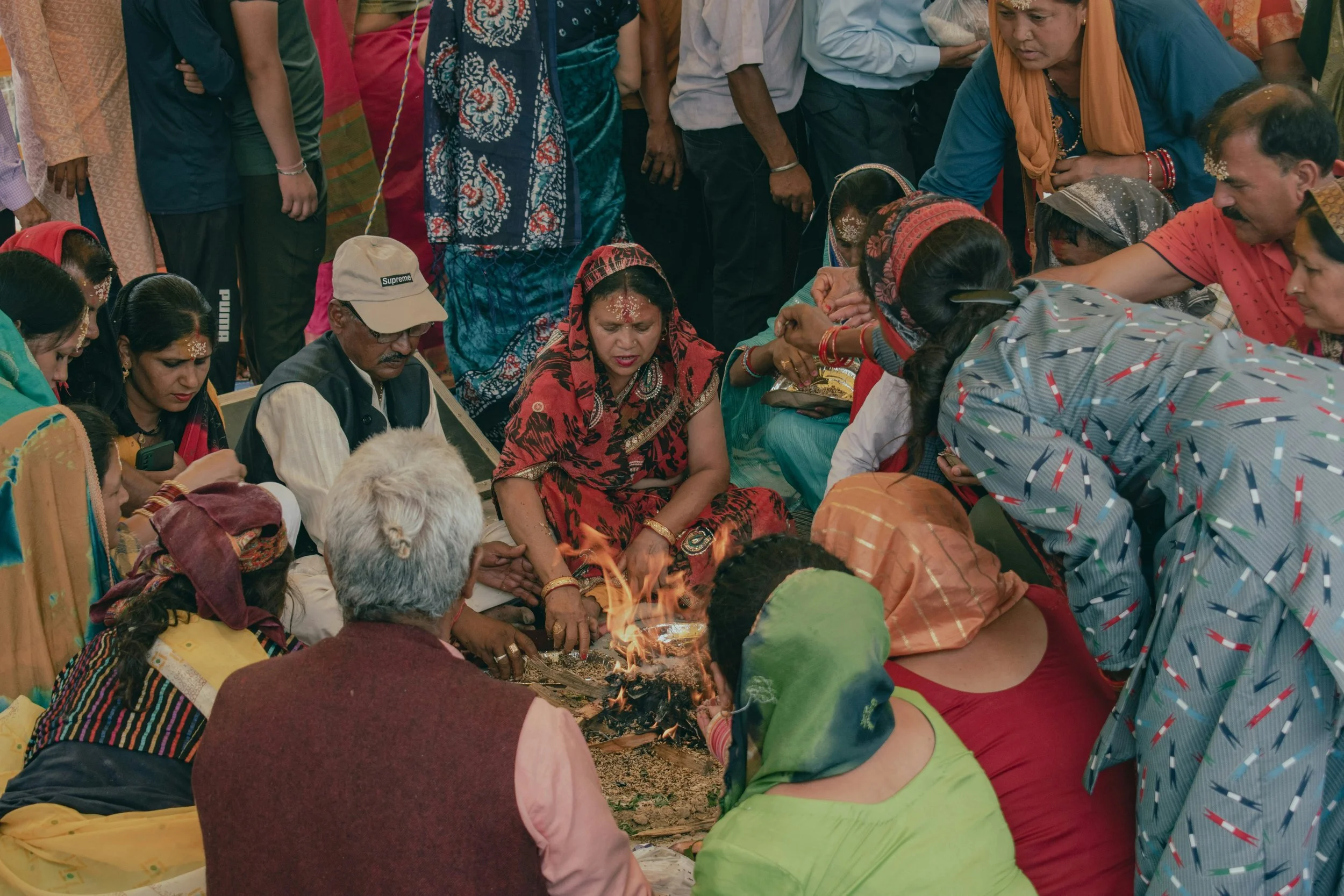 Group of people gathered around a sacred fire, participating in a traditional Hindu ceremony, with some women wearing colorful traditional clothing and decorative markings on their foreheads.