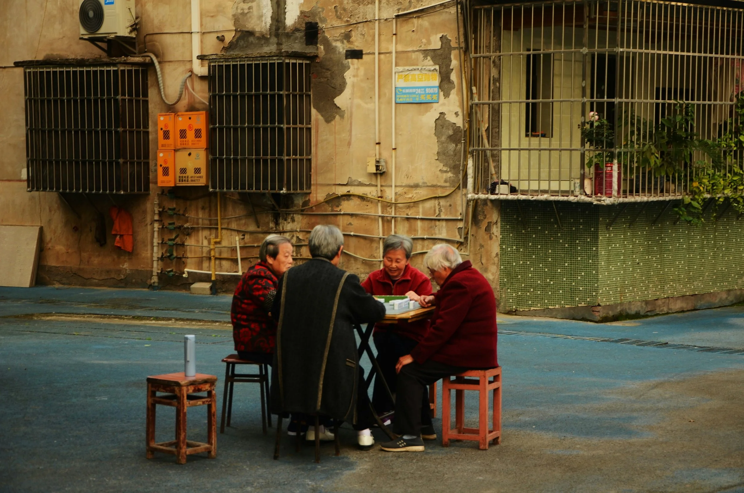 Four elderly people sitting around a table playing a game or cards on a street in an urban area, with old buildings in the background.