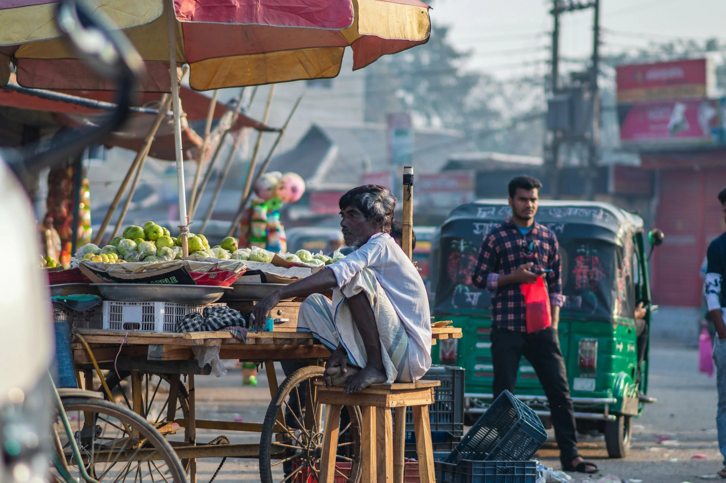 An outdoor street market scene with a middle-aged man sitting behind a wooden cart filled with green apples. The man is wearing a white shirt and plaid lungi. Several colorful umbrellas shade the market stalls. In the background, there are pedestrians, auto rickshaws, and buildings with signage, suggesting a busy urban area in India.