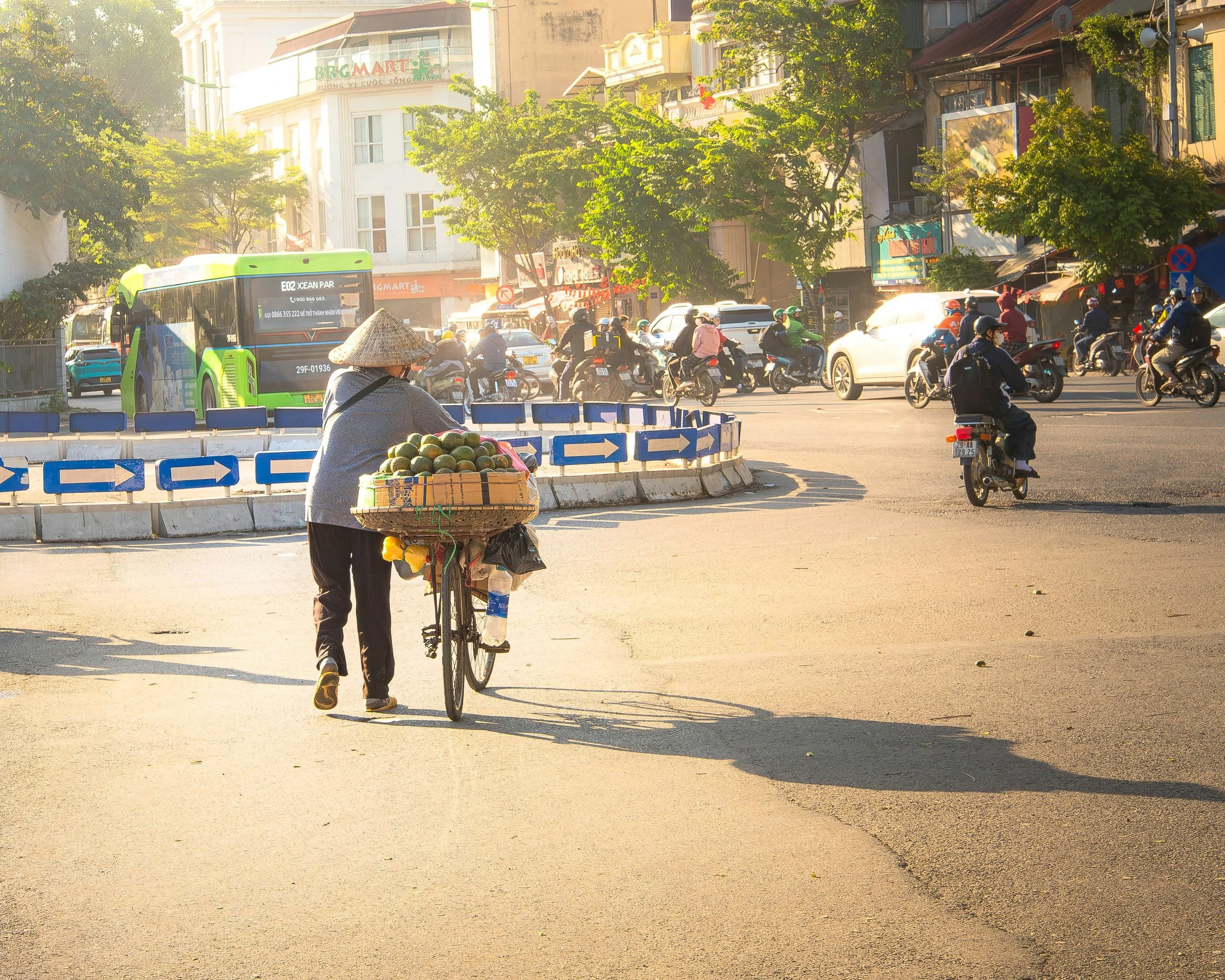 A busy city intersection with a person wearing traditional Asian conical hat selling fruits from a basket on a bicycle, surrounded by motorbikes and cars, with buildings, trees, and a bus in the background, in warm sunlight.