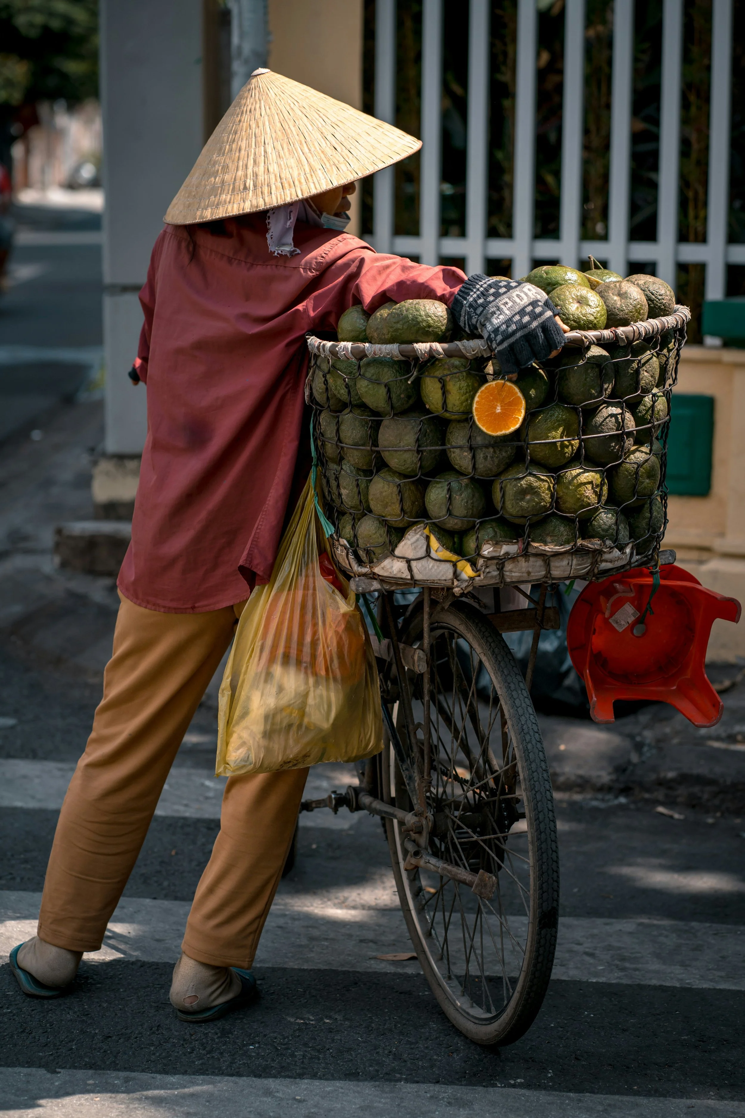 A person wearing a straw hat and a pink jacket, pushing a bicycle with a large basket filled with avocados and a pulled orange inside, on a street.