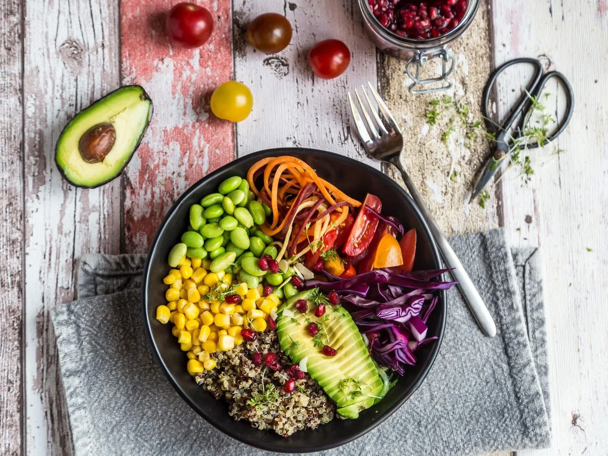 A black bowl filled with a colorful salad containing avocado slices, red cabbage, cherry tomatoes, corn, and quinoa, on a rustic wooden table with a fork placed inside the bowl, alongside a sliced avocado, cherry tomatoes, scissors, and a jar of pomegranate seeds.