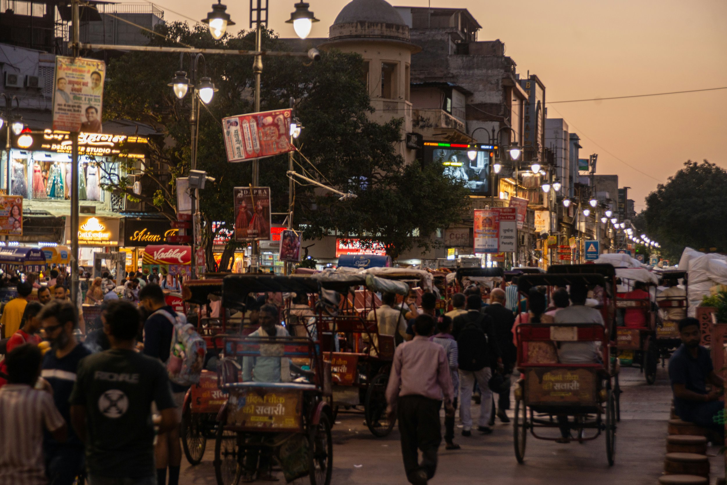 A busy street market scene during sunset with many people, street vendors, and rickshaws. Buildings with illuminated signs and streetlights are visible in the background.