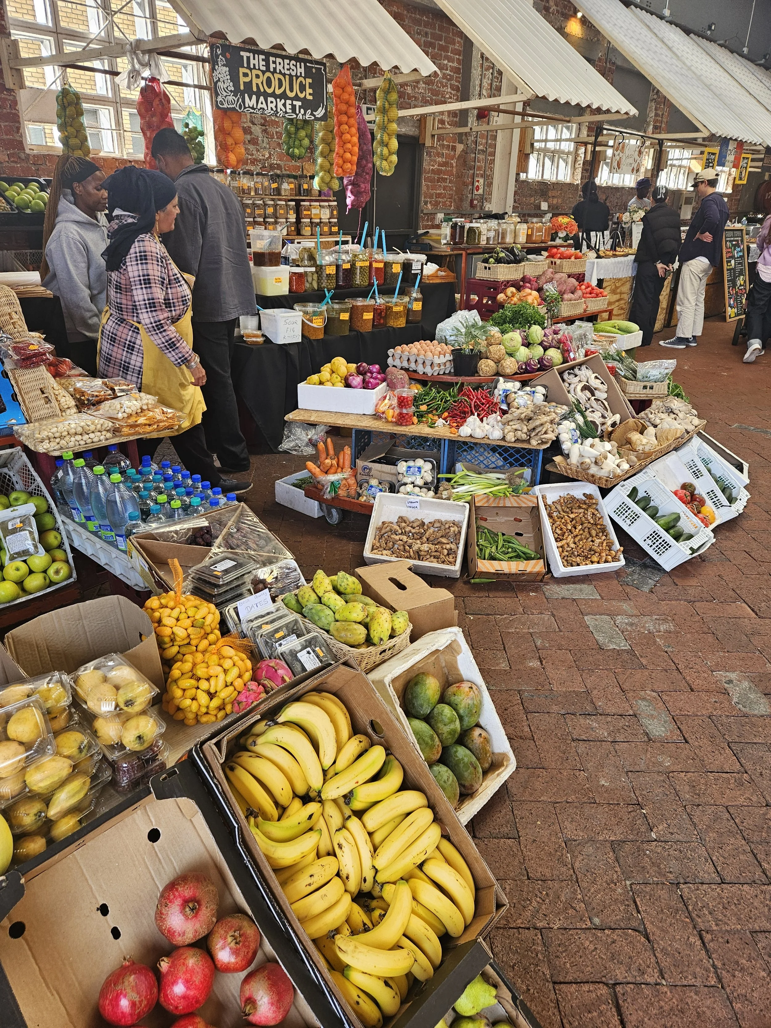 A busy market stall with fresh produce including apples, bananas, pomegranates, and various vegetables. Several customers are browsing, and jars of condiments or preserves are displayed along with hanging citrus and onions. The stall has a sign that reads 'The Fresh Produce Market'.