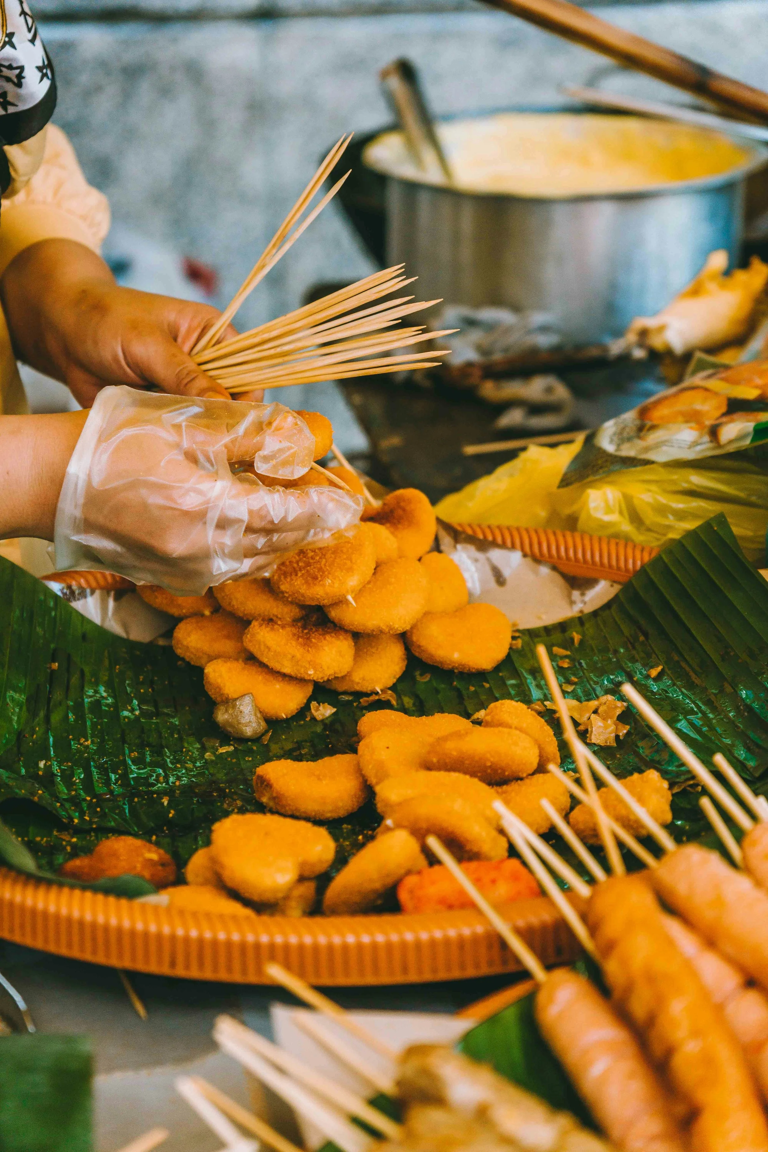 Close-up of skewered fried foods on a bamboo tray at a street food stall, with a stainless steel pot in the background.