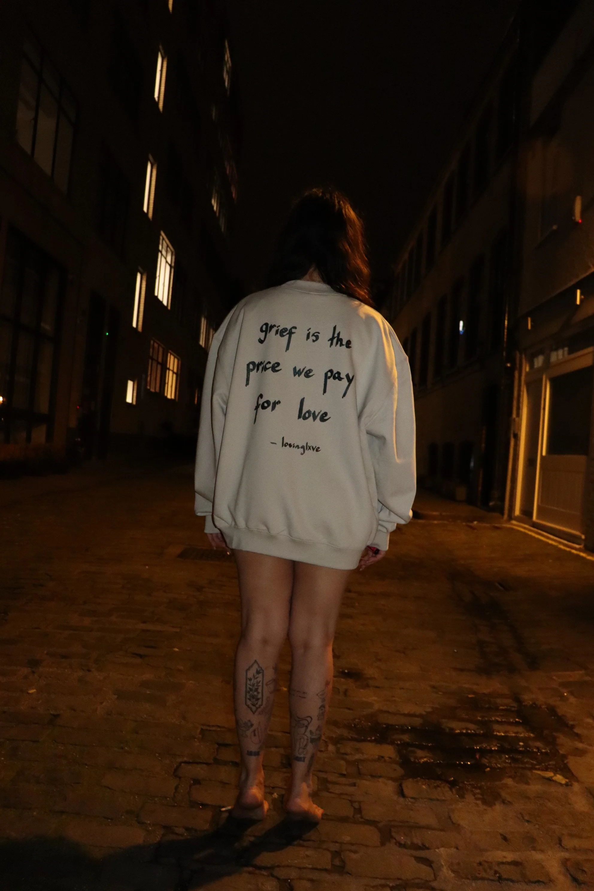 Person walking at night on cobblestone street, wearing an oversized white sweatshirt with text on the back, and showing tattoos on the legs.