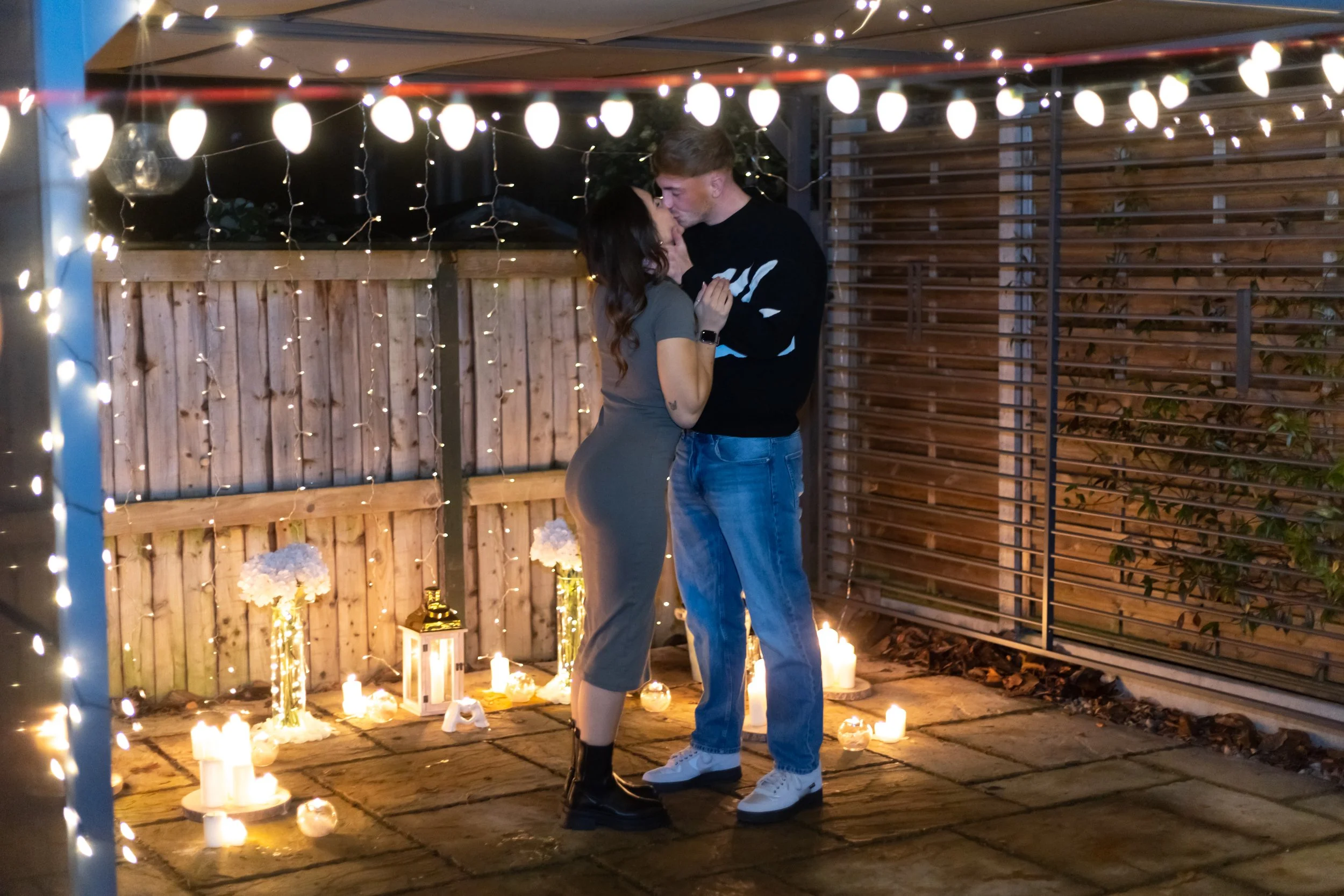 A couple is sharing a kiss in an outdoor setting decorated with string lights, candles, and lanterns, indicating a romantic moment.