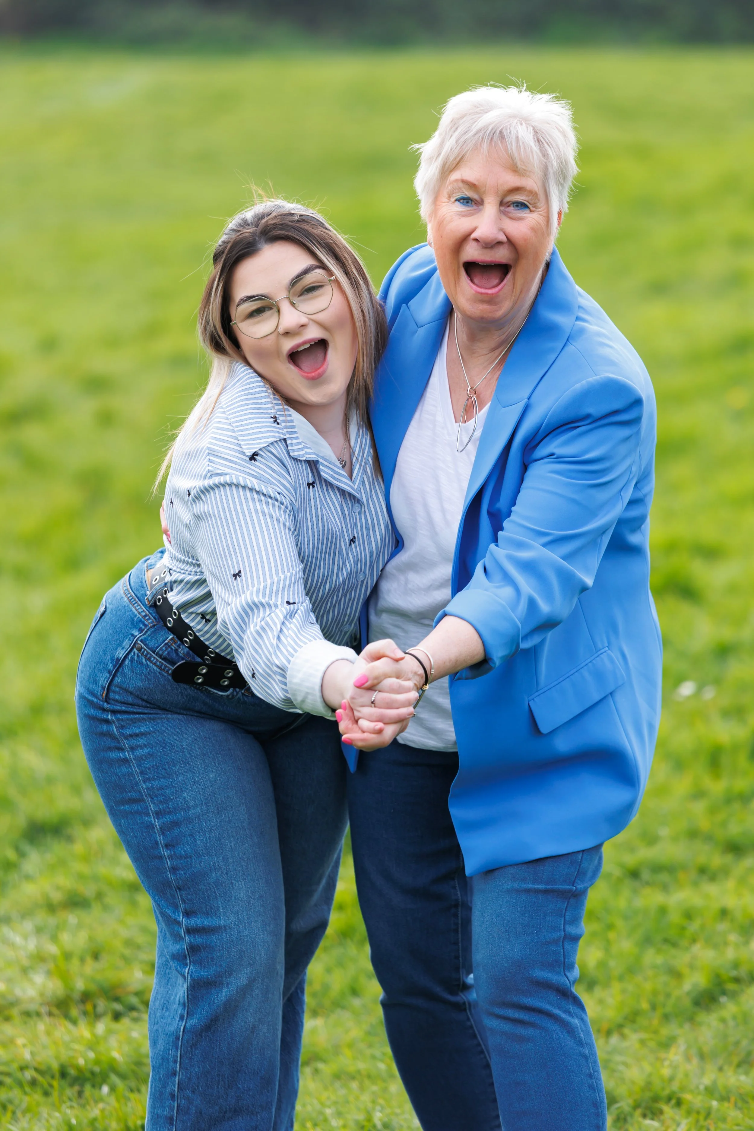 An elderly woman and a young woman are holding hands and laughing together outdoors on a grassy field. The elderly woman has short white hair, is wearing a bright blue blazer over a white shirt, and has a necklace. The young woman has long dark hair,