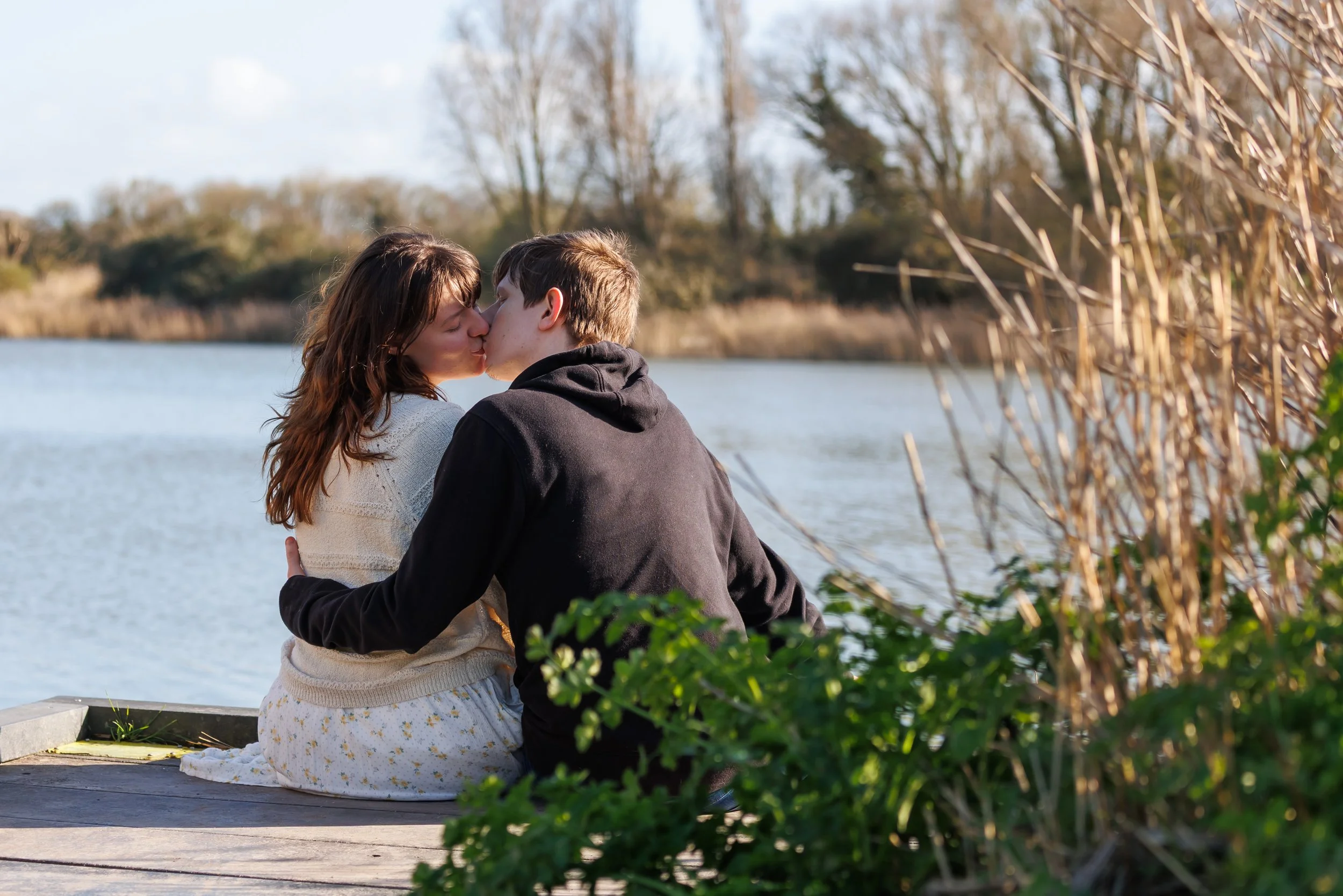 A couple shares a kiss outdoors near a body of water, surrounded by trees and tall grasses, with a cloudy sky in the background.