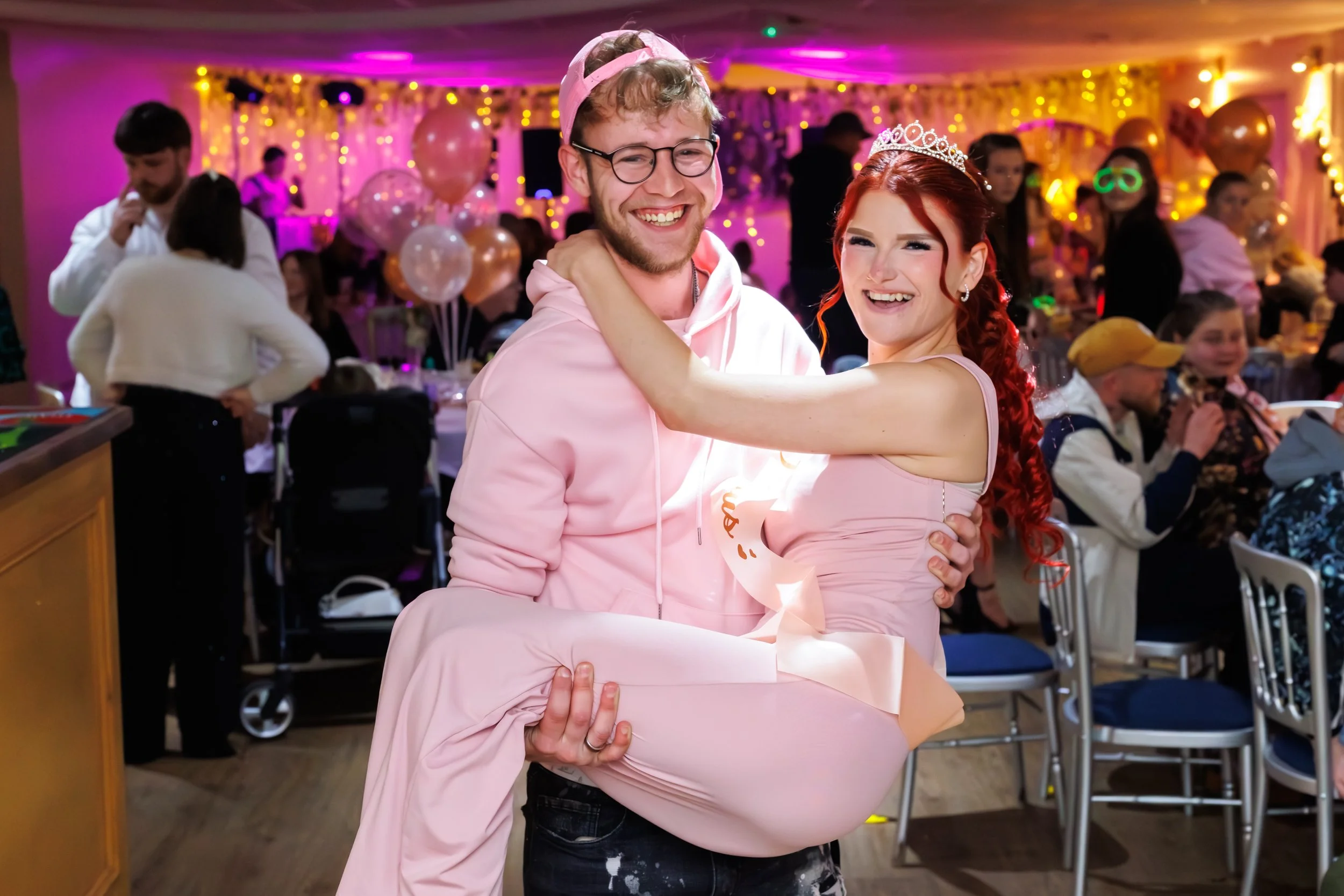 A man and woman celebrating at a party, wearing pink outfits and tiaras, smiling and dancing in a decorated hall with pink and gold balloons and string lights.
