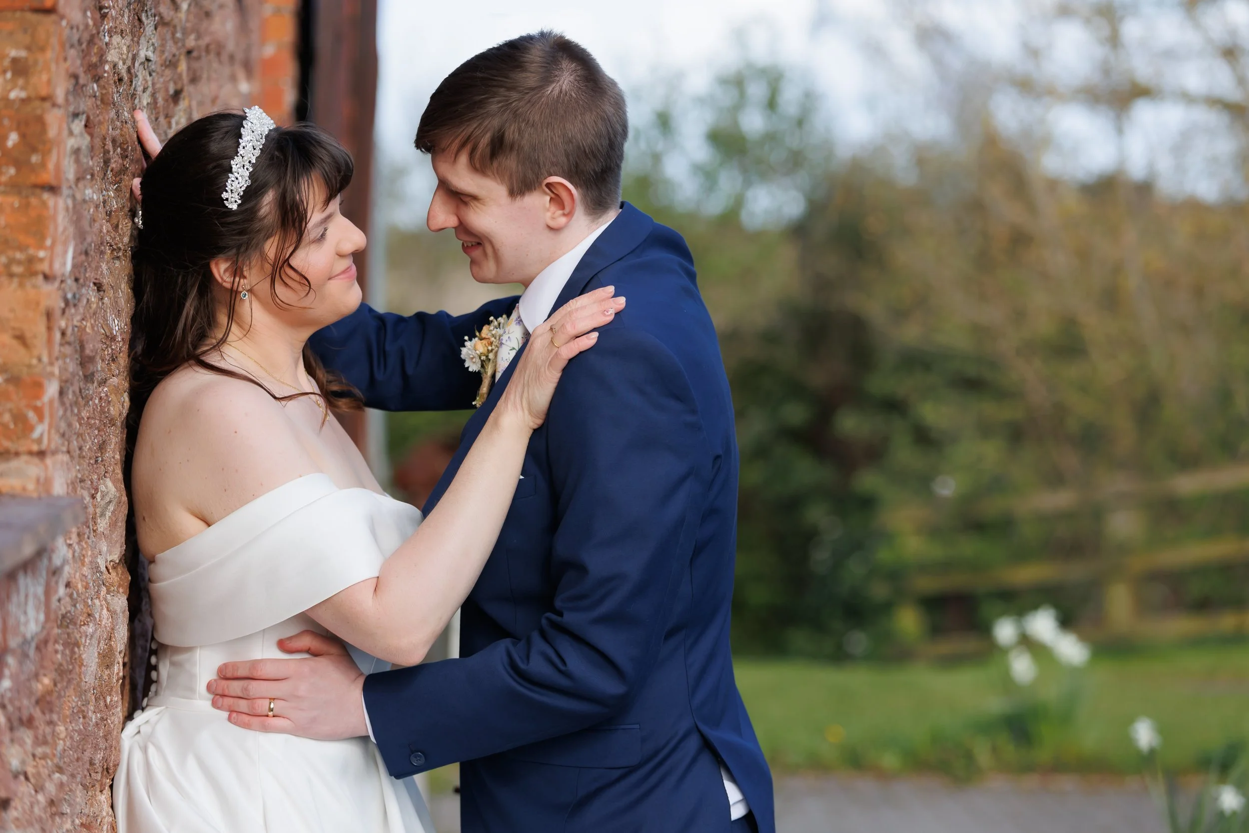 A bride and groom, facing each other with their foreheads and noses almost touching, standing close to a brick wall outdoors. The bride has dark hair and is wearing a white off-the-shoulder dress with a sparkling headband. The groom is wearing a blue
