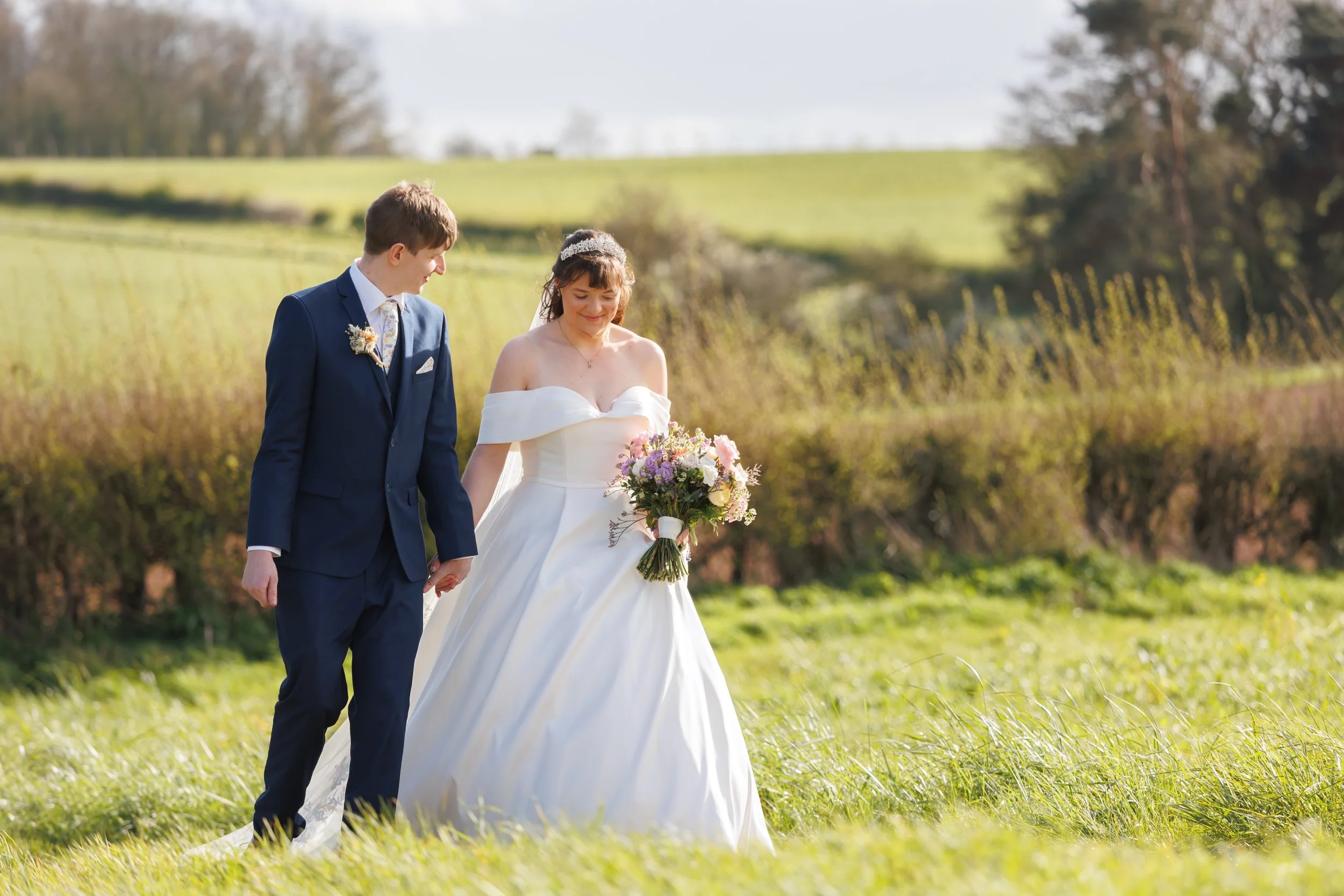 A bride and groom walking hand in hand through a grassy field outdoors during their wedding, with the bride holding a bouquet of flowers.