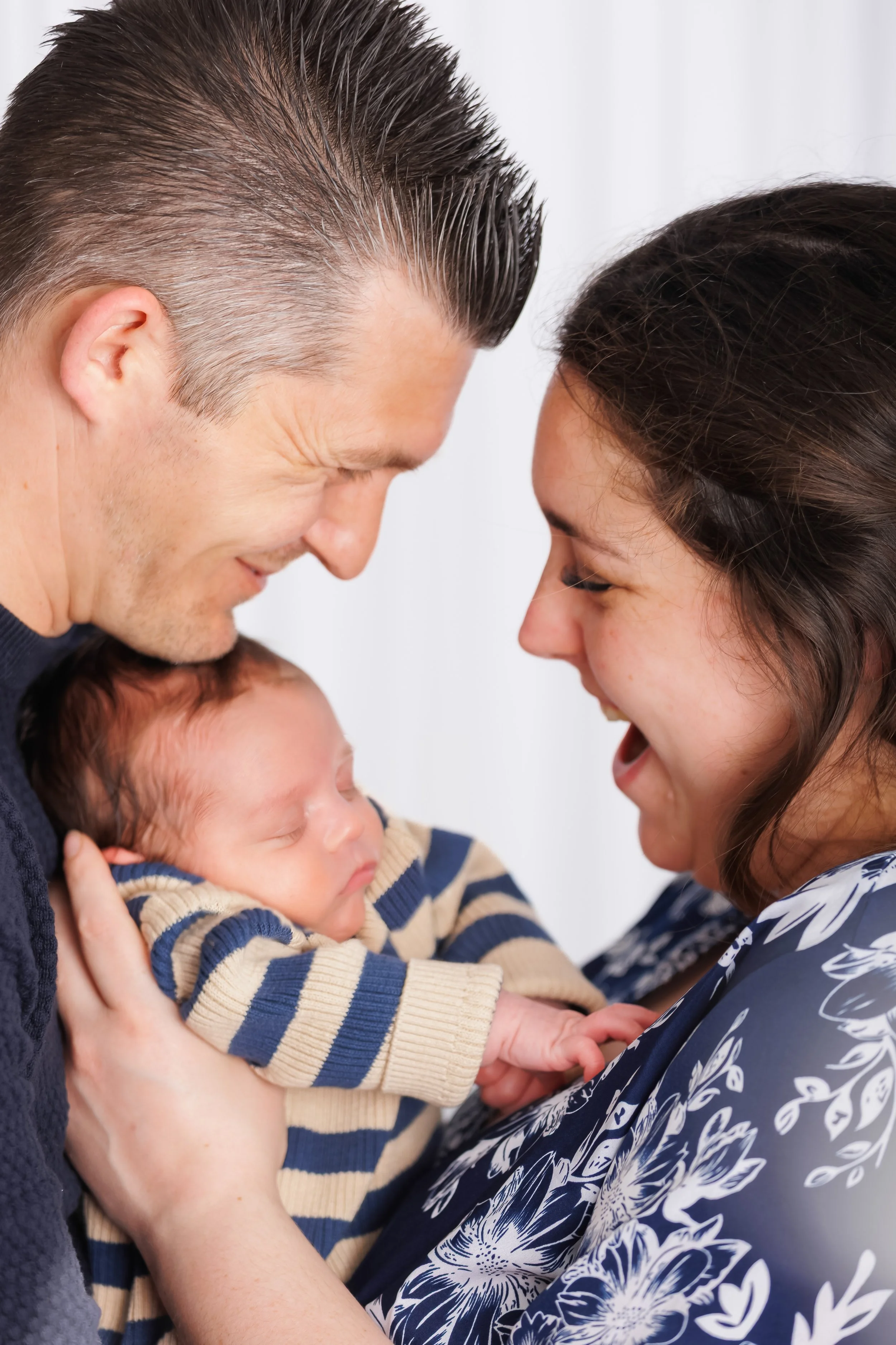 A man, woman, and baby are close together, with the man and woman touching foreheads and smiling while the baby sleeps in the woman's arms.